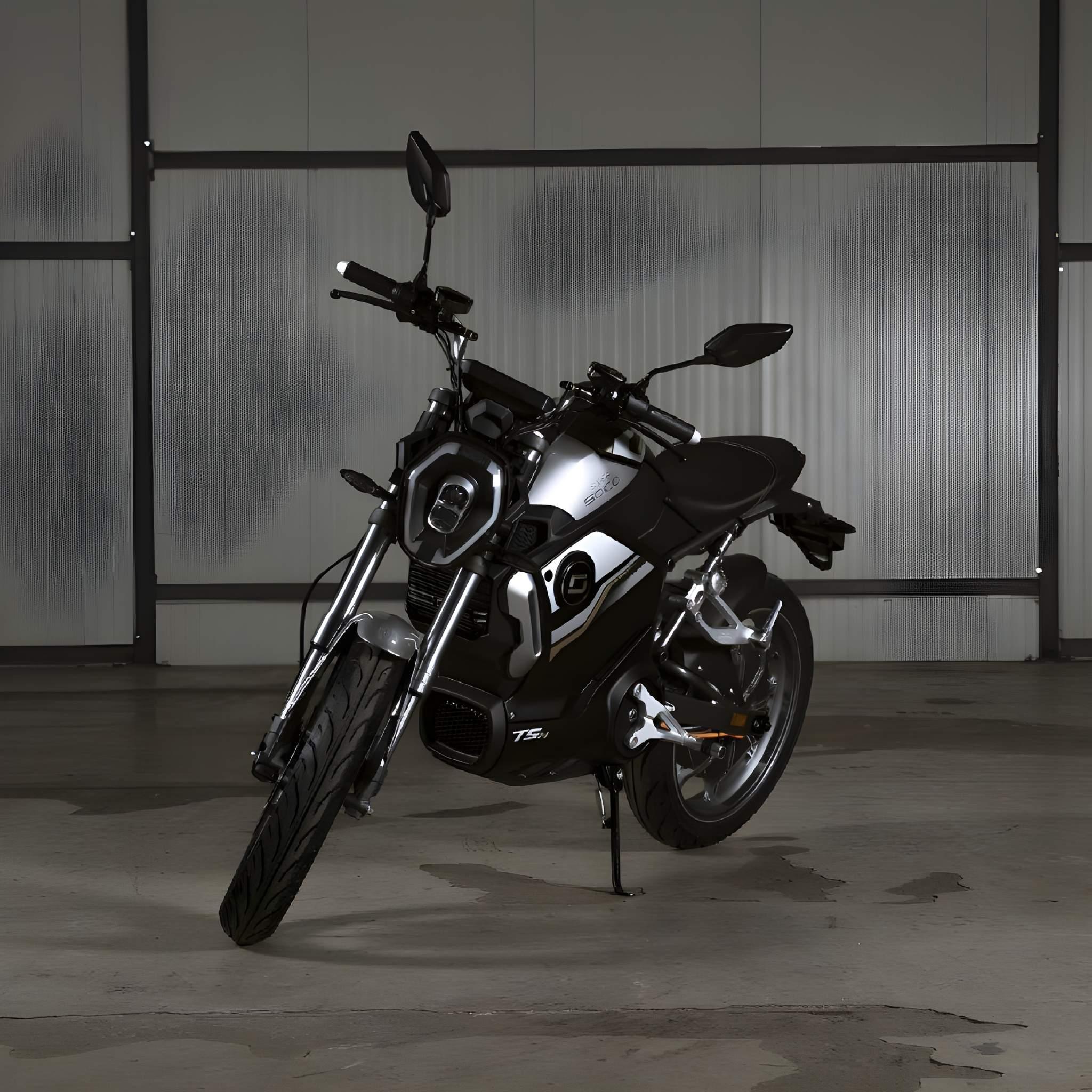 Ein elegantes schwarz-silbernes Super Soco E-Motorrad TSx Eco 45 km/h steht auf einem Betonboden vor Metall- und Glaspaneelen und präsentiert modernes Design und emissionsfreie Mobilität.