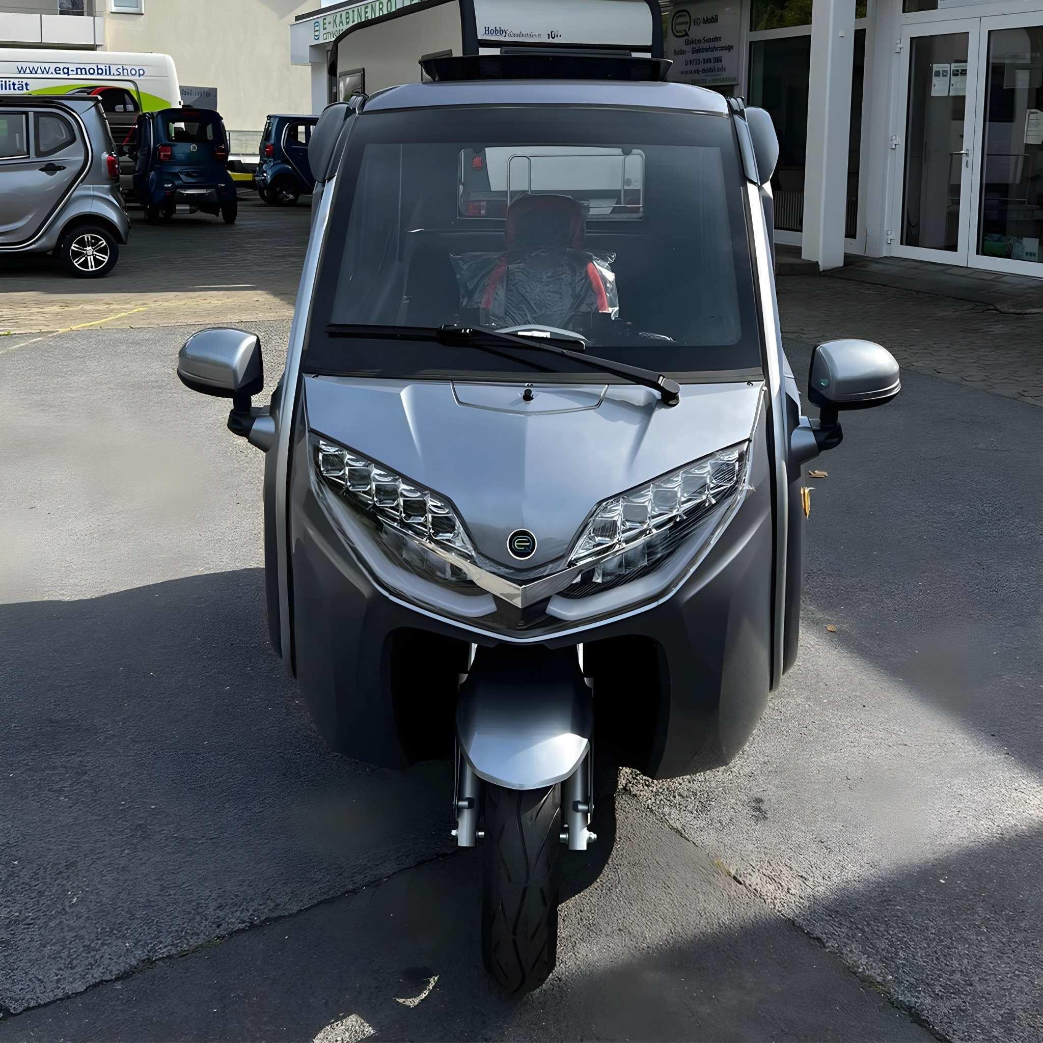 Frontansicht eines EQ-Mobil Kabinenroller Lima Q3 Li 45 / 25 km/h (aktuelles Modell) mit Lithium-Akku, ein geschlossenes dreirädriges Elektrofahrzeug, geparkt auf einem gepflasterten Parkplatz mit anderen Fahrzeugen und Gebäuden im Hintergrund.