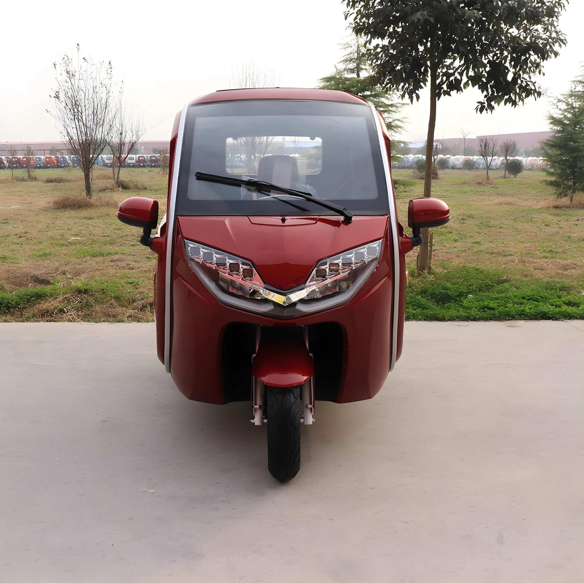 Ein EQ-Mobil Kabinenroller Lima Q3 Li 45 / 25 km/h mit einem Lithium-Akku, in rot, ist im Freien auf Beton geparkt, mit Blick nach vorne. Im Hintergrund sind Grasflächen und Bäume zu sehen. Es ist ein dreirädriges geschlossenes Elektrofahrzeug.