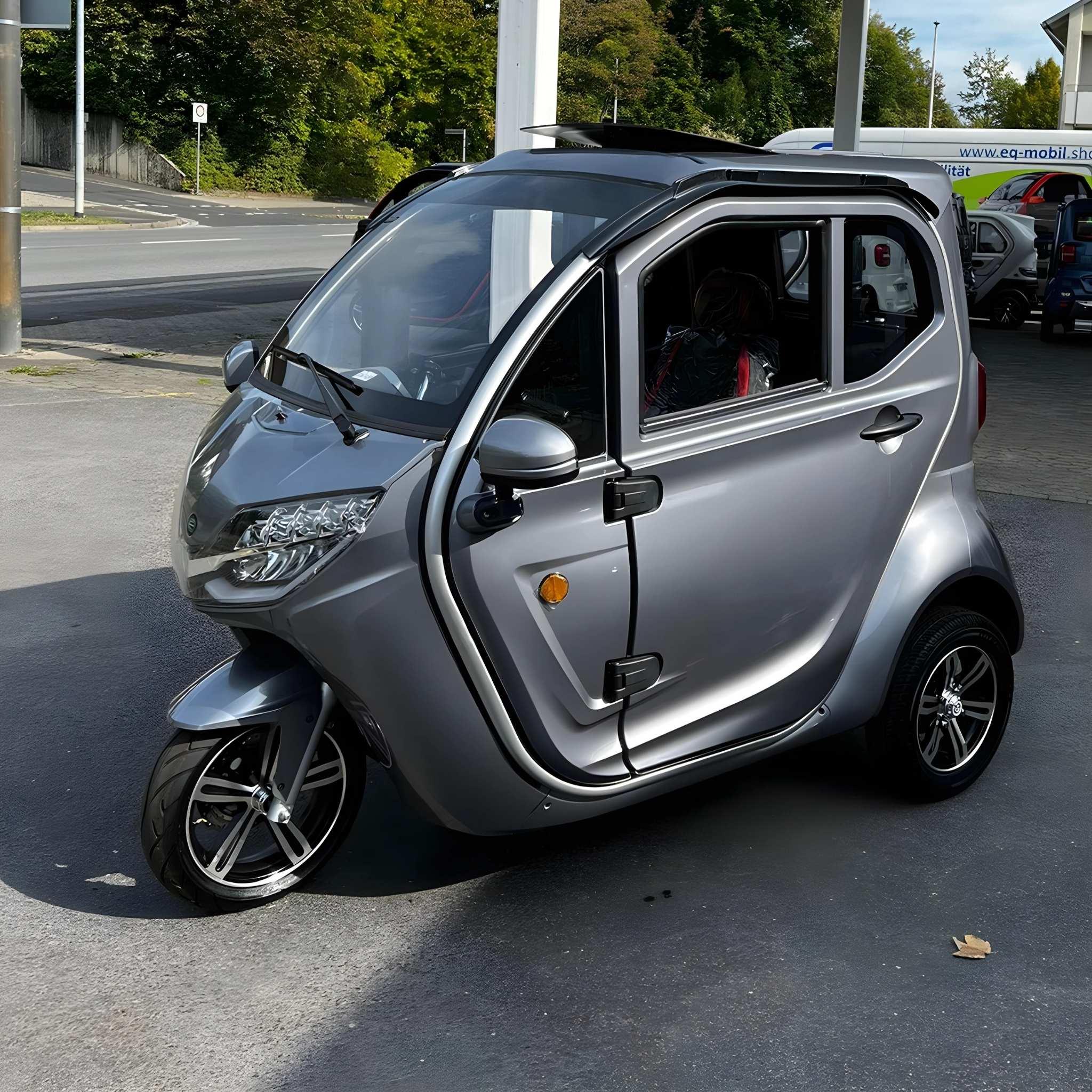 Ein kompakter, silberner EQ-Mobil Kabinenroller Lima Q3 Li 45/25 km/h mit Lithium-Batterie parkt auf einer gepflasterten Fläche inmitten von Grünflächen und Straßen im Hintergrund.