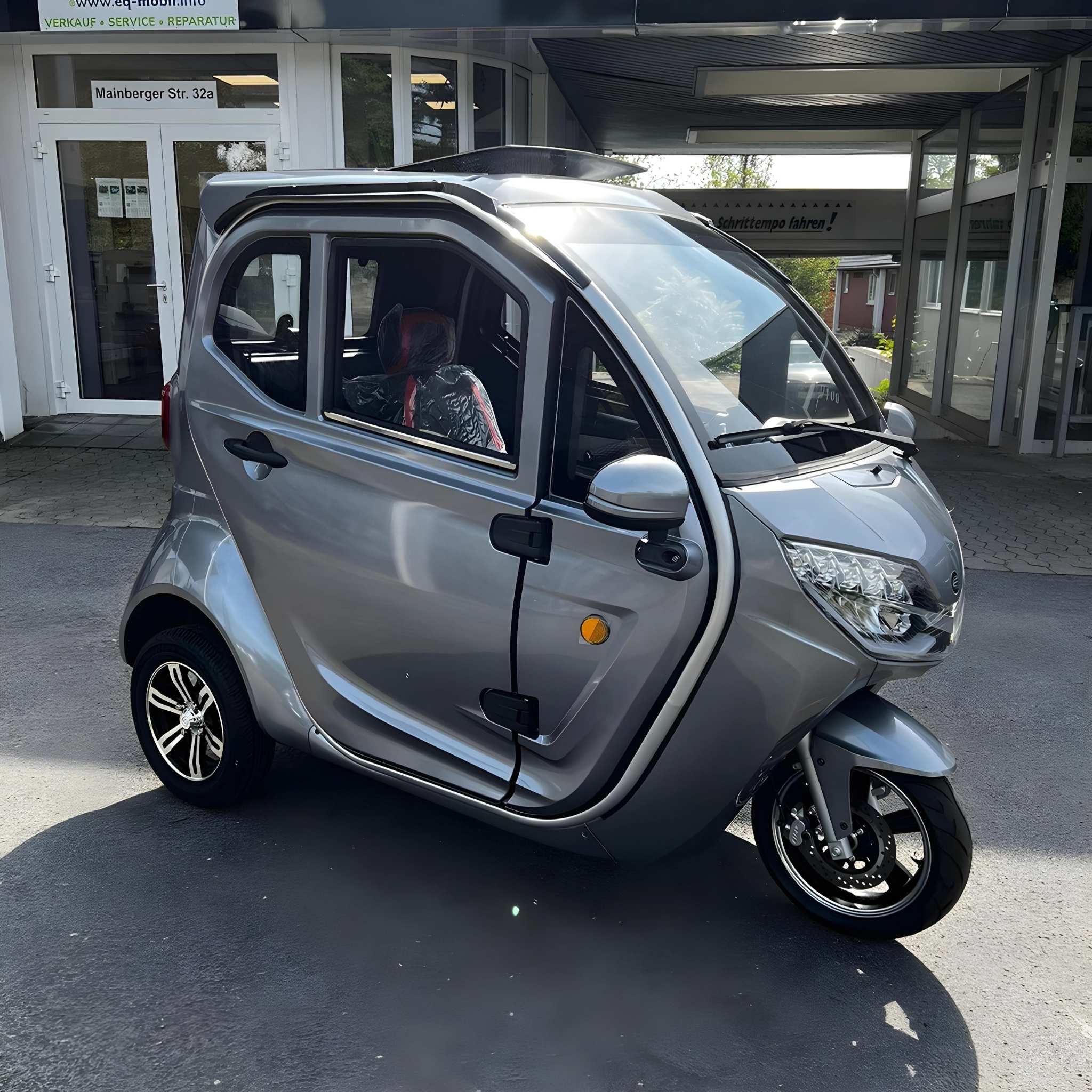 Ein kompakter, silberner EQ-Mobil Kabinenroller Lima Q3 Li 45 / 25 km/h mit Lithium-Akku, drei Rädern, zwei Türen und schwarzen Fenster- und Radzierblenden ist an einem sonnigen Tag vor einem Gebäude geparkt.