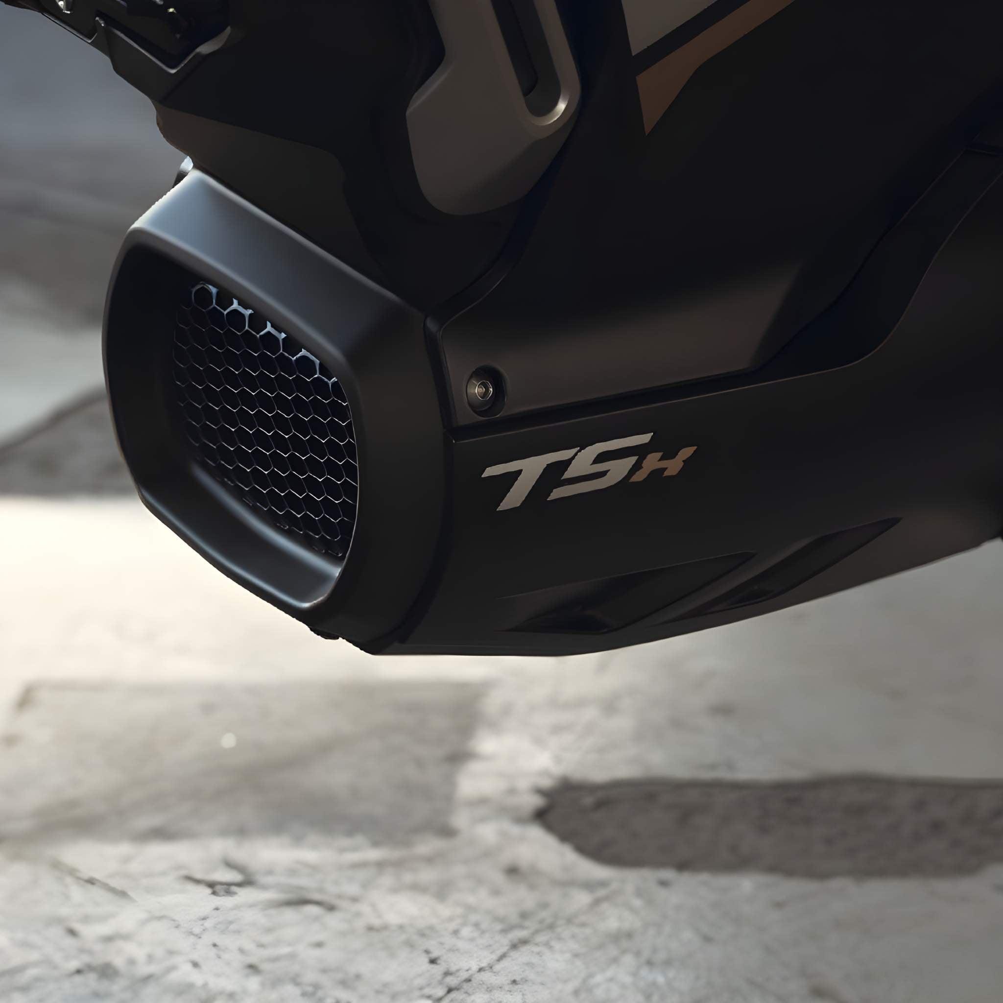 Nahaufnahme des E-Motorrads SUPER SOCO TSx Eco 45 km/h von Super Soco mit Wabengrill und TSH-Logo auf dem schwarzen Auspuff. Der sanft verschwommene Betonhintergrund unterstreicht das emissionsfreie Design.