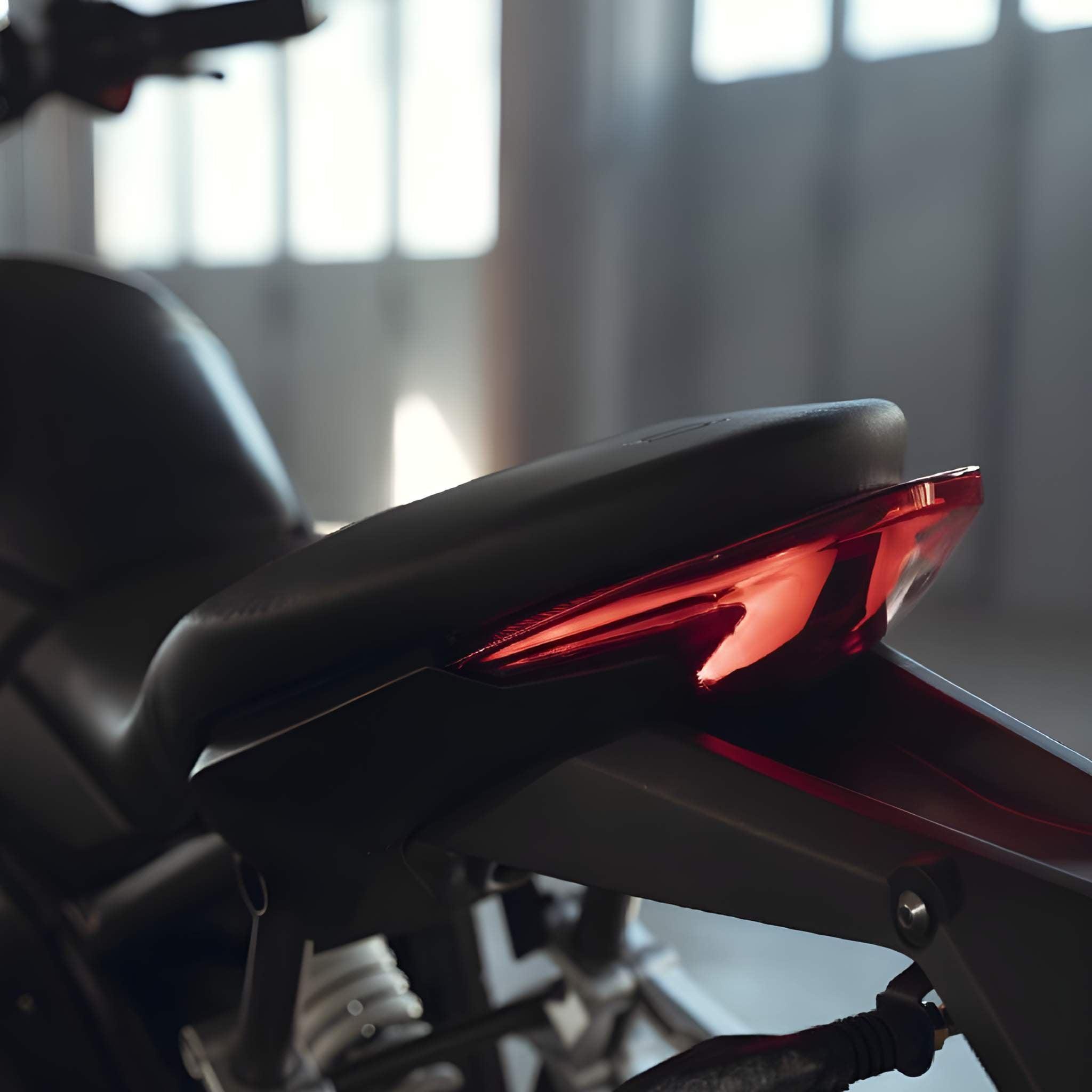 Nahaufnahme der hinteren Sitzbank und des roten Rücklichts des Super Soco E-Motorrad TSx Eco 45 km/h, wobei die glatte schwarze Sitzbank vor einem sanft verschwommenen Innenhintergrund mit natürlichem Licht zu sehen ist.