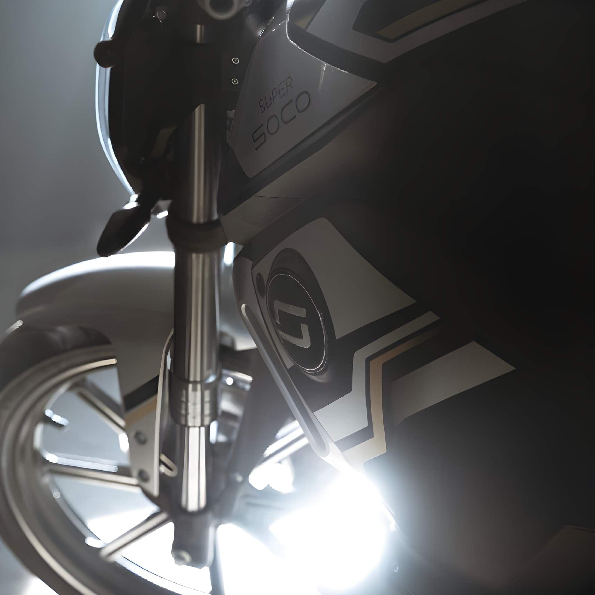 Nahaufnahme des Vorderrads, der Gabel und der Karosserie des Super Soco E-Motorrad TSx Eco 45 km/h, mit geometrischem Logo und heller Hintergrundbeleuchtung.