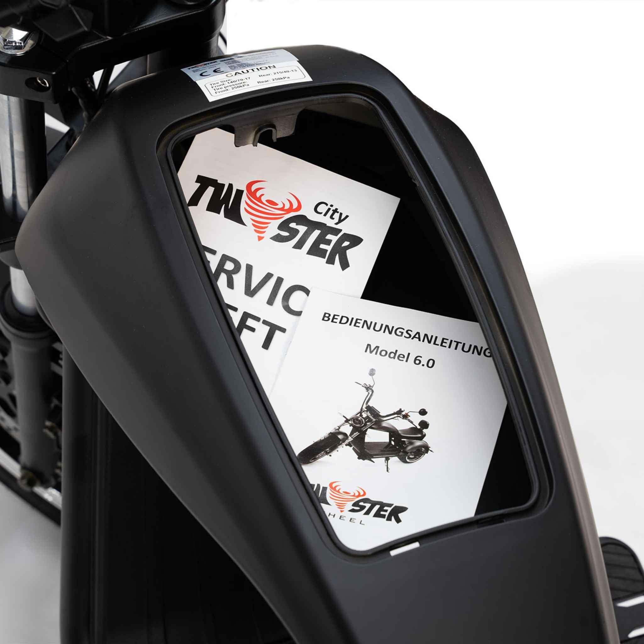Eine Nahaufnahme des Staufachs eines schwarzen City Twister E-Choppers zeigt zwei Bedienungsanleitungen: eine mit der Aufschrift City Twister" und einer Grafik sowie eine weitere in deutscher Sprache für das CityTwister 6.0 MAX DUO 80 km/h Carplay Display Modell 2026.