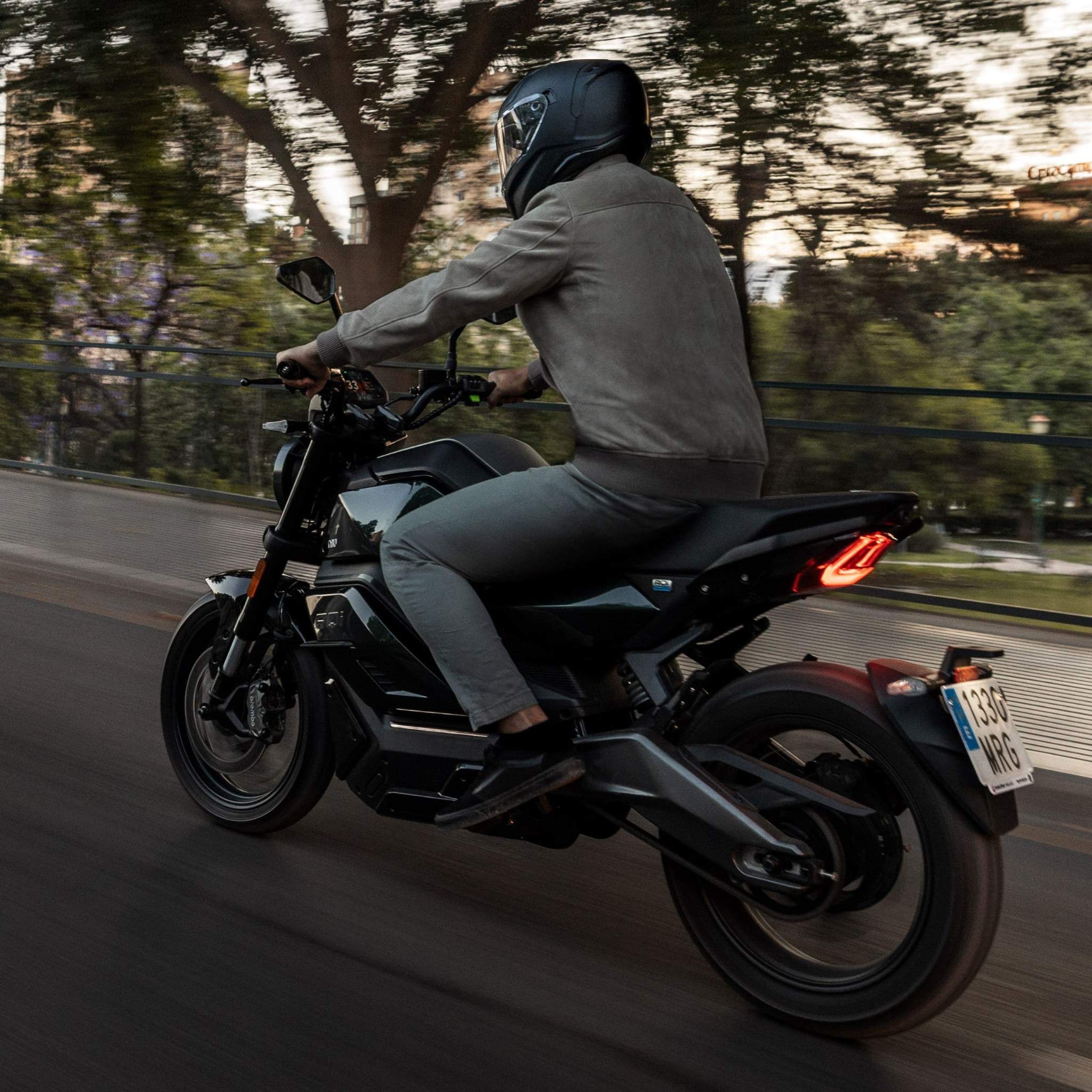 Ein behelmter Fahrer in einer Jacke rast auf dem NIU Elektromotorrad RQI Sport (110 km/h) durch die Straßen der Stadt, wobei Bäume und Gebäude im Hintergrund verschwimmen und ein Gefühl der Bewegung eingefangen wird.