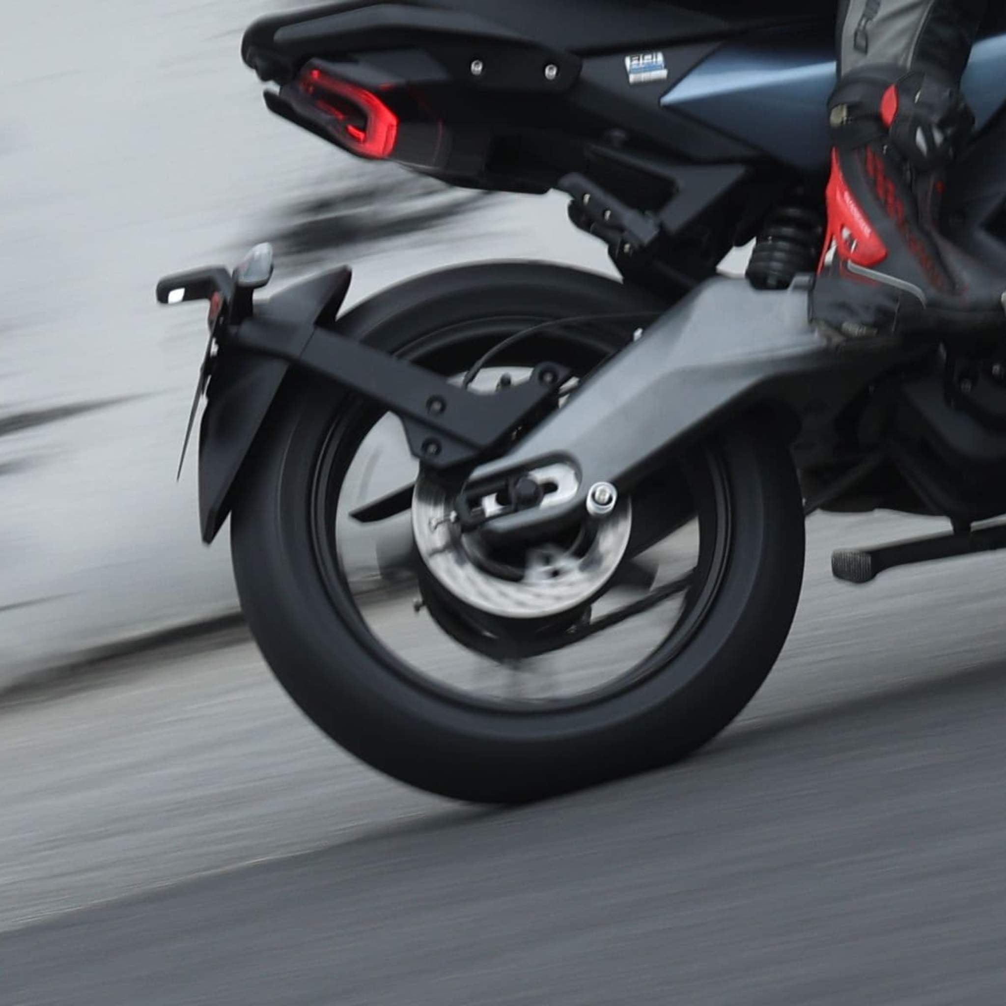 Nahaufnahme des Hinterrads des NIU Elektromotorrad RQI Sport 110 km/h, kein Auspuff sichtbar, gefahren von einer Person mit roten und schwarzen Stiefeln. Das Fahrrad ist in Bewegung und neigt sich nach links auf die Straße.