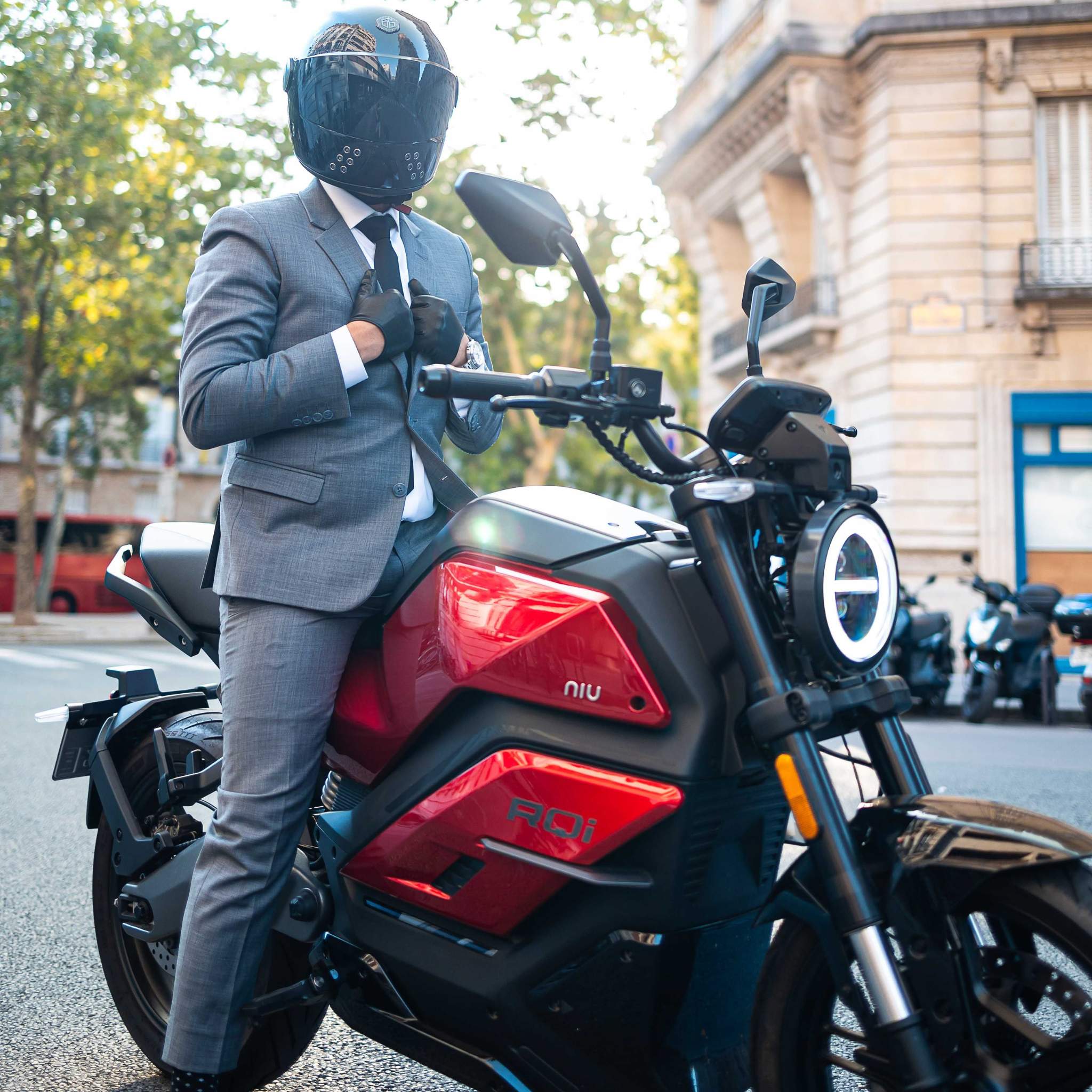 Eine Person im grauen Anzug und mit schwarzem Helm sitzt selbstbewusst auf einem rot-schwarzen NIU Elektromotorrad RQI Sport 110 km/h, das auf einer Stadtstraße mit Bäumen und klassischen Gebäuden im Hintergrund geparkt ist.