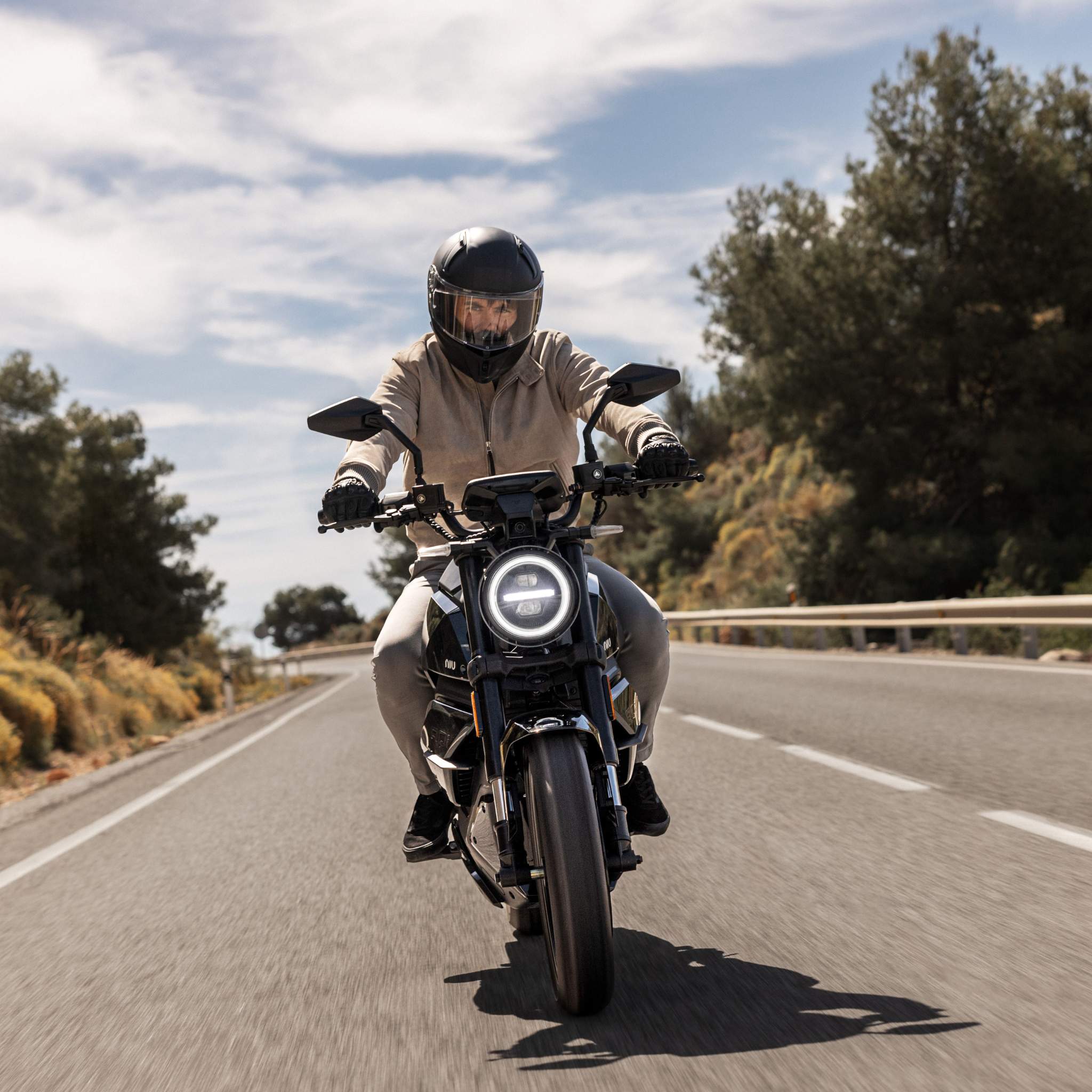 Ein Fahrer mit Helm und leichter Kleidung fährt auf einem schnittigen schwarzen NIU Elektromotorrad RQI Sport 110 km/h auf einer sonnigen, von Bäumen gesäumten Straße mit Leitplanken unter einem teilweise bewölkten Himmel.