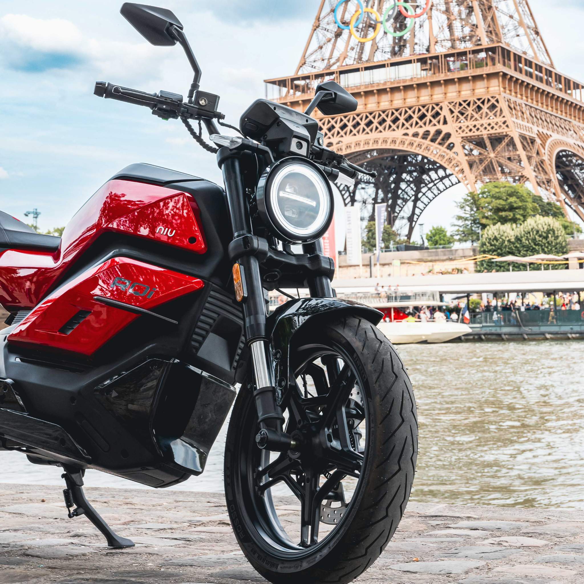 Ein rotes NIU Elektromotorrad RQI Sport 110 km/h ist auf einem Kopfsteinpflasterweg an der Seine geparkt. Im Hintergrund ist der Eiffelturm zu sehen, der Himmel ist teilweise bewölkt.