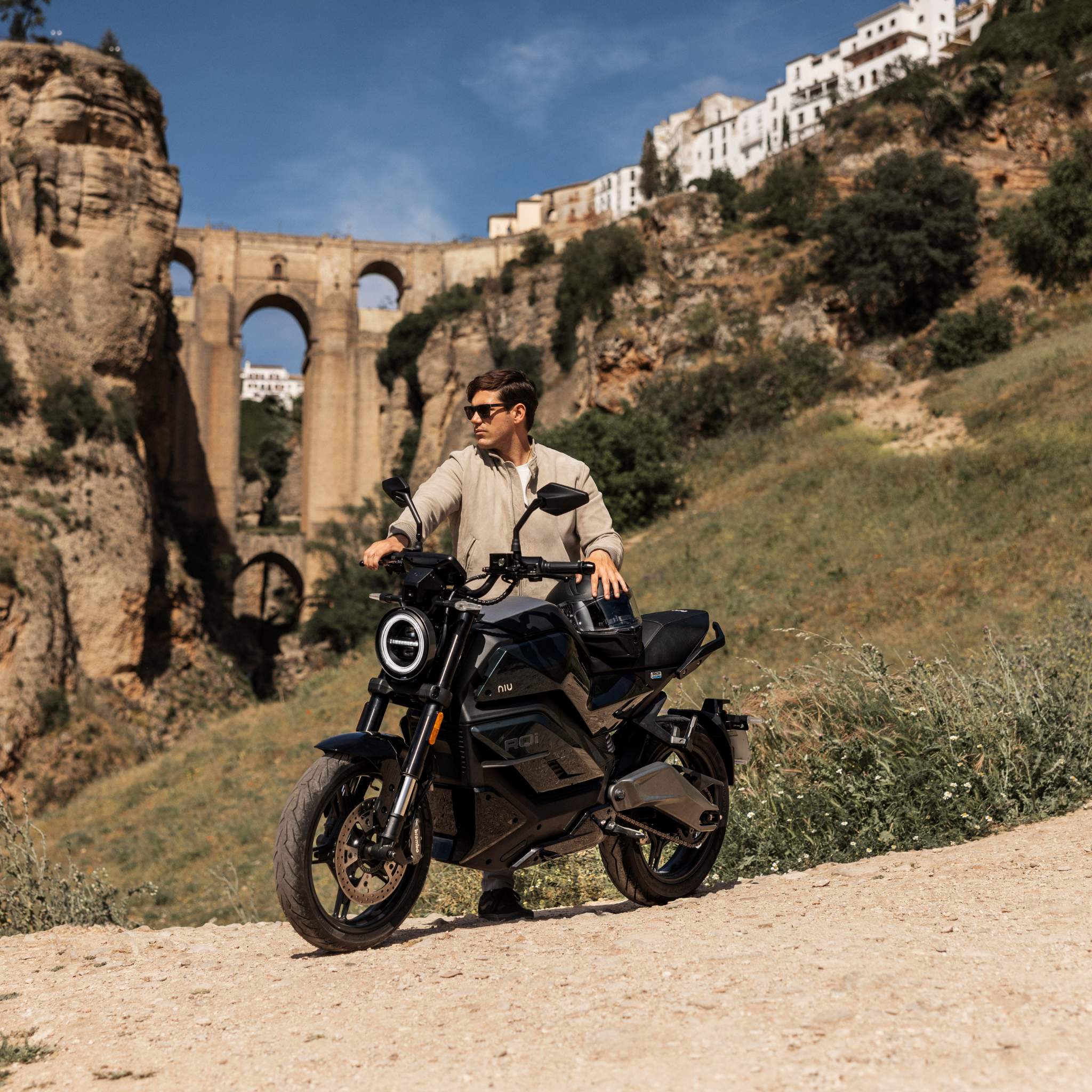 Eine Person mit Sonnenbrille und beiger Kleidung steht neben einem NIU Elektromotorrad RQI Sport 110 km/h auf einem unbefestigten Weg, mit einer historischen Steinbrücke und weißen Gebäuden in den Hügeln im sonnigen Hintergrund.