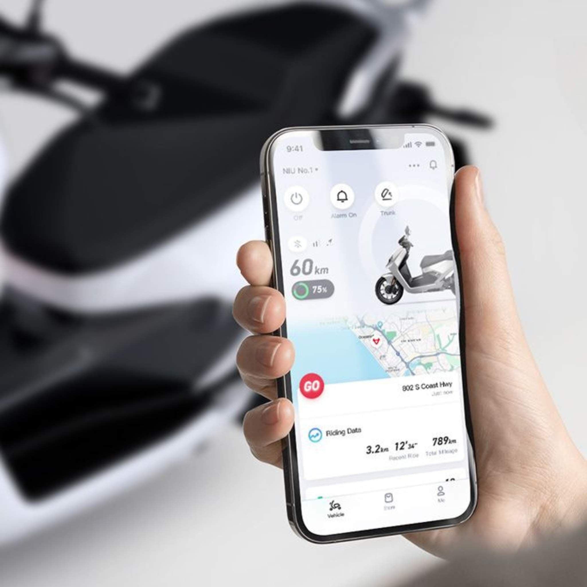 Eine Hand hält ein Smartphone, auf dem die NIU FQI 500 App mit Fahrdaten, Routenkarte, Geschwindigkeit, Batteriestand und Scooterbild angezeigt wird. Im Hintergrund ist ein verschwommener NIU Elektroroller E-Scooter Motorroller FQI 500 (95 km/h) - Neues Modell 2025.