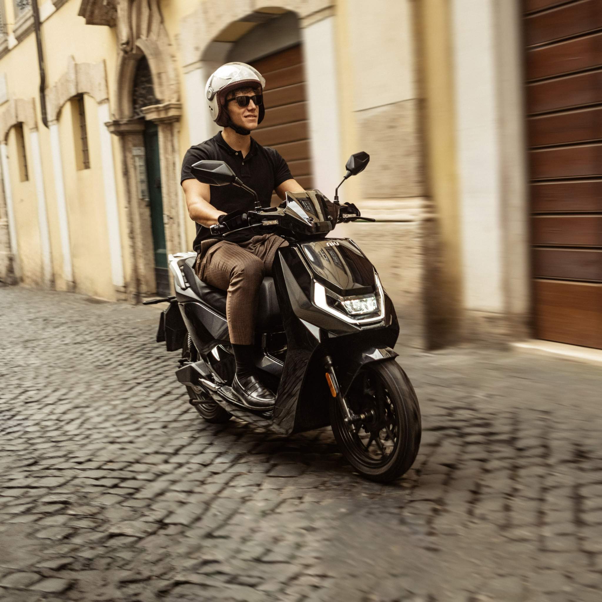 Eine Person mit Helm und Sonnenbrille fährt mit dem NIU Elektroroller Motorroller FQI 500 (Modell 2025, 95 km/h) auf einem schwarzen Roller über eine Kopfsteinpflasterstraße in einer historischen Gegend mit alten Gebäuden im Hintergrund.