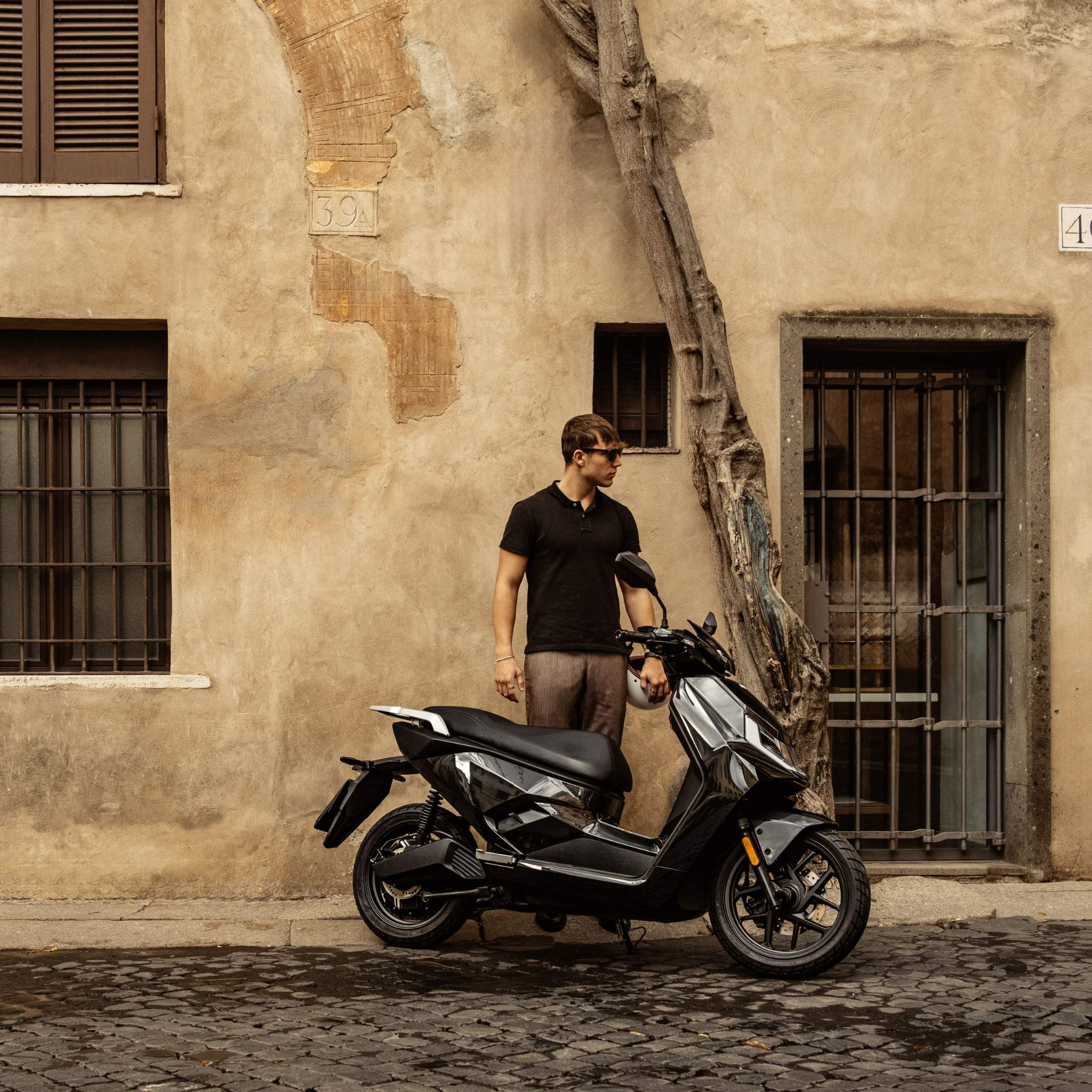 Ein Mann mit Sonnenbrille und schwarzem Hemd steht neben dem stylischen NIU Elektroroller Motorroller FQI 500 (Modell 2025, 95 km/h) auf einer Kopfsteinpflasterstraße vor einem alten beigen Gebäude mit vergitterten Fenstern und einem Baum an der Wand.