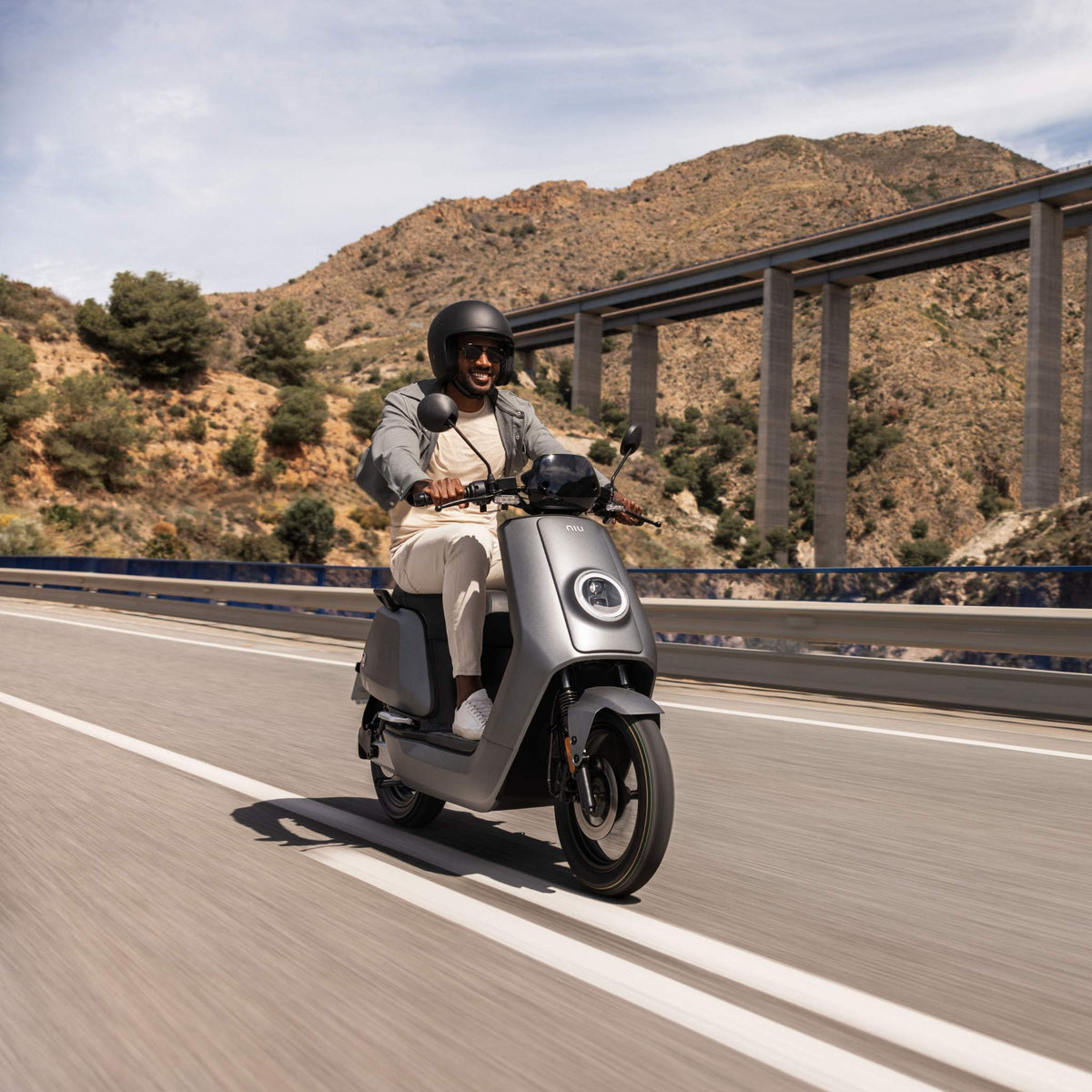 Eine Person mit Helm und Sonnenbrille fährt mit dem stylischen NIU Elektroroller Motorroller NQI GTS (2024 Upgrade) mit 45 km/h auf einer Autobahn mit Bergen und einer Brücke und genießt Elektromobilität unter dem sonnigen Himmel.
