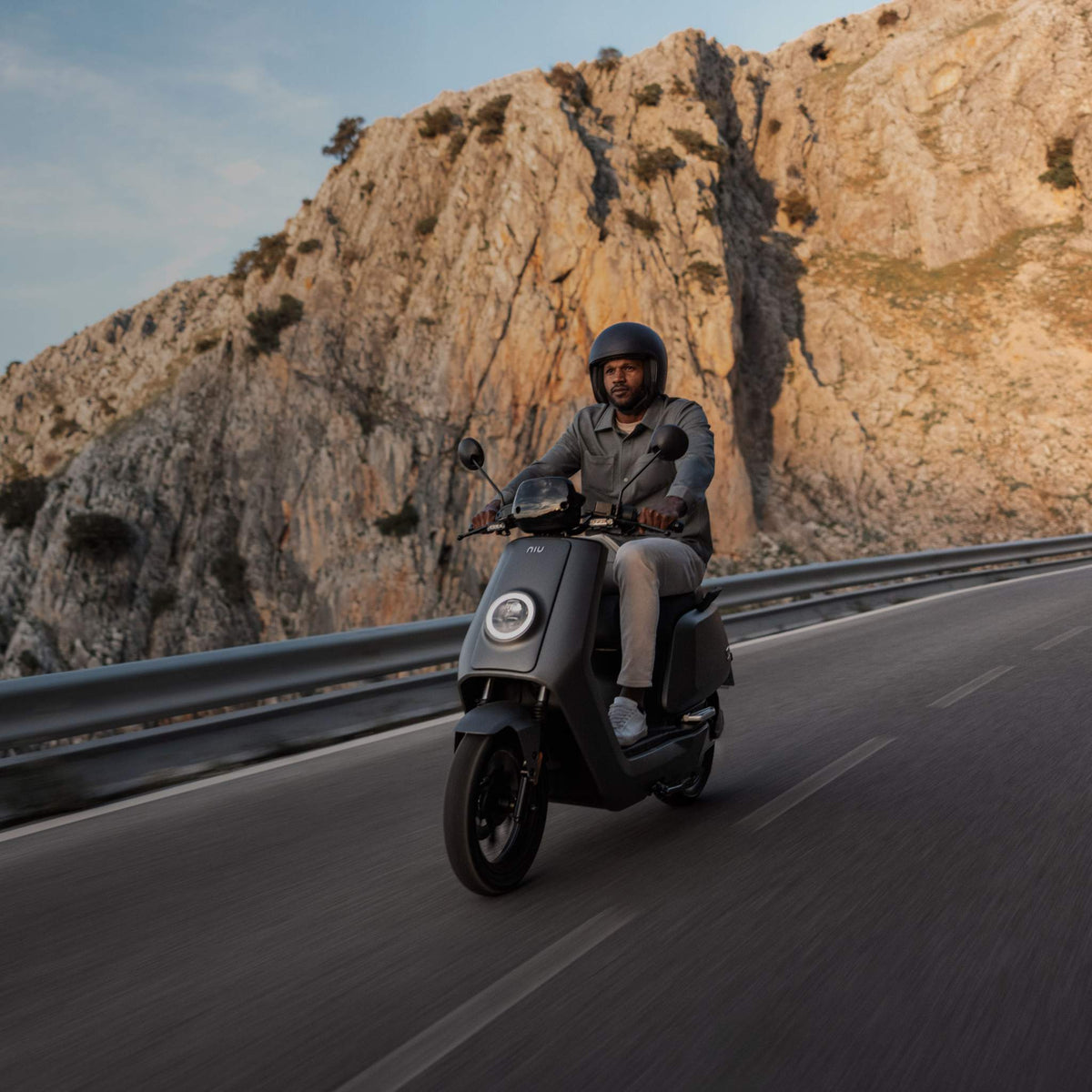 Eine Person mit Helm fährt mit dem schwarzen NIU Elektroroller Motorroller NQI GTS (2024 Upgrade, 45 km/h) auf einer Bergstraße mit felsigen Klippen in der Abendsonne - Elektromobilität in atemberaubender Kulisse genießen.