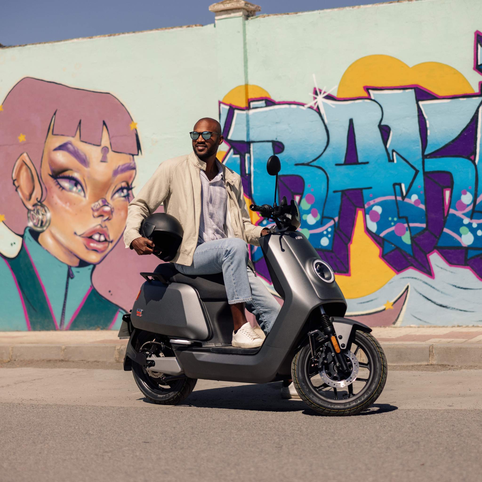 Ein Mann mit Sonnenbrille und heller Jacke sitzt auf einem grauen NIU Elektroroller Motorroller NQI GTS (2024 Upgrade) 45 km/h und hält einen Helm. Er parkt neben einem Wandbild mit stilisiertem Frauengesicht und plakativem Graffiti, das Elektromobilität umarmt.
