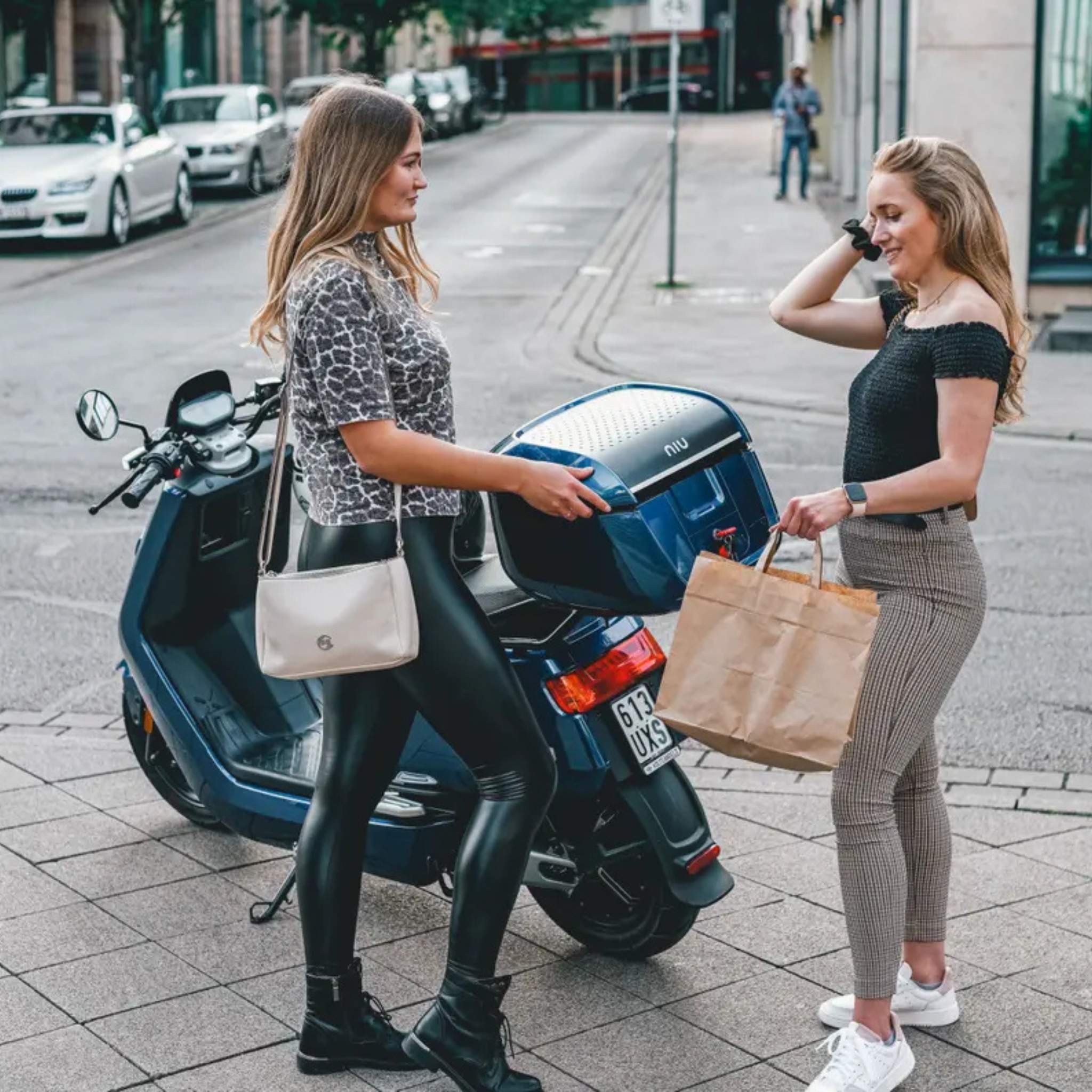 Zwei Frauen stehen neben einem blauen NIU Elektroroller E-Scooter Motorroller NQI Sport (2024 Upgrade) 45 km/h auf einem städtischen Bürgersteig. Die eine hält Einkaufstüten in der Hand und lächelt, die andere, in schwarzen Stiefeln und Kunstlederhosen, steht mit einer Handtasche daneben.