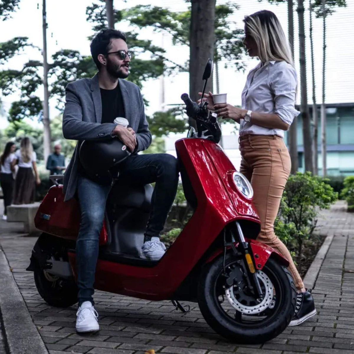 Ein Mann sitzt auf einem roten NIU Elektroroller E-Scooter Motorroller NQI Sport (2024 Upgrade) 45 km/h, hält einen Helm und einen Kaffee in der Hand und unterhält sich im Freien mit einer Frau, die mit einer Tasse in der Nähe steht, inmitten von Bäumen und Grünflächen.