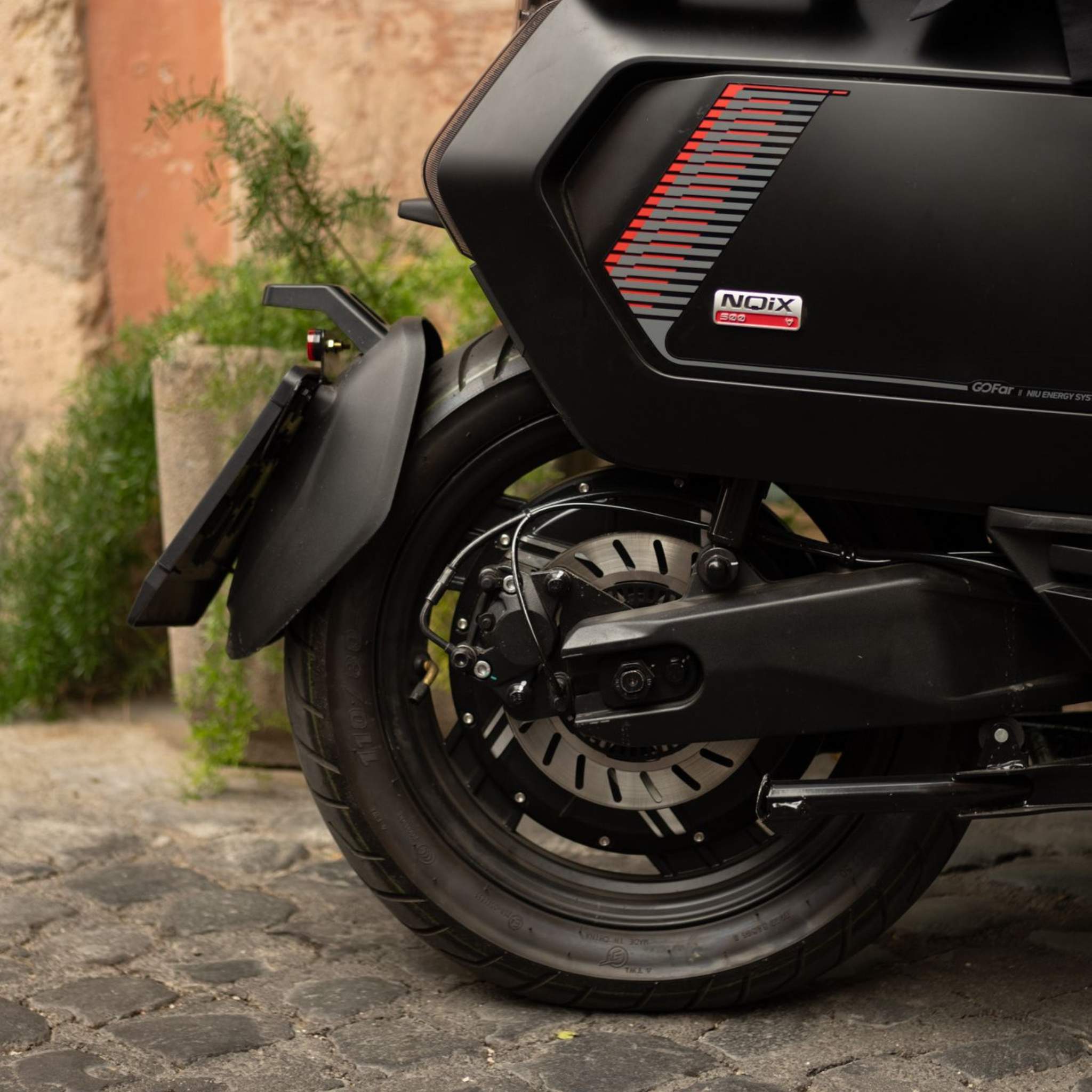 Nahaufnahme des Hinterrads und des Kotflügels eines schwarzen NIU Elektrorollers E-Scooter Motorroller NQiX 500 (100 km/h) auf Kopfsteinpflaster, mit Pflanzgefäß und Mauer im Hintergrund. Rote Streifenaufkleber und NQiX-Branding auf dem NIU Elektroroller sichtbar.
