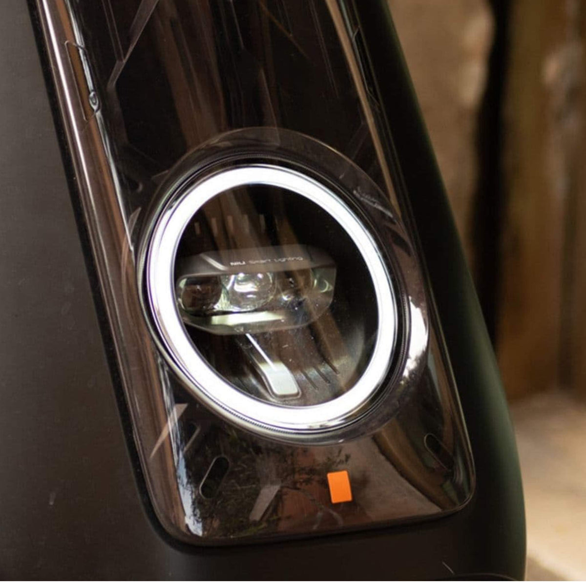 Nahaufnahme des Scheinwerfers am NIU NQiX 500 Elektroroller E-Scooter Motorroller Modell 2025 mit einem kreisförmigen LED-Ring und einer klaren Linse, die auf einer glänzend schwarzen Oberfläche montiert ist. Unterhalb des Scheinwerfers ist ein kleines orangefarbenes Quadrat zu sehen.