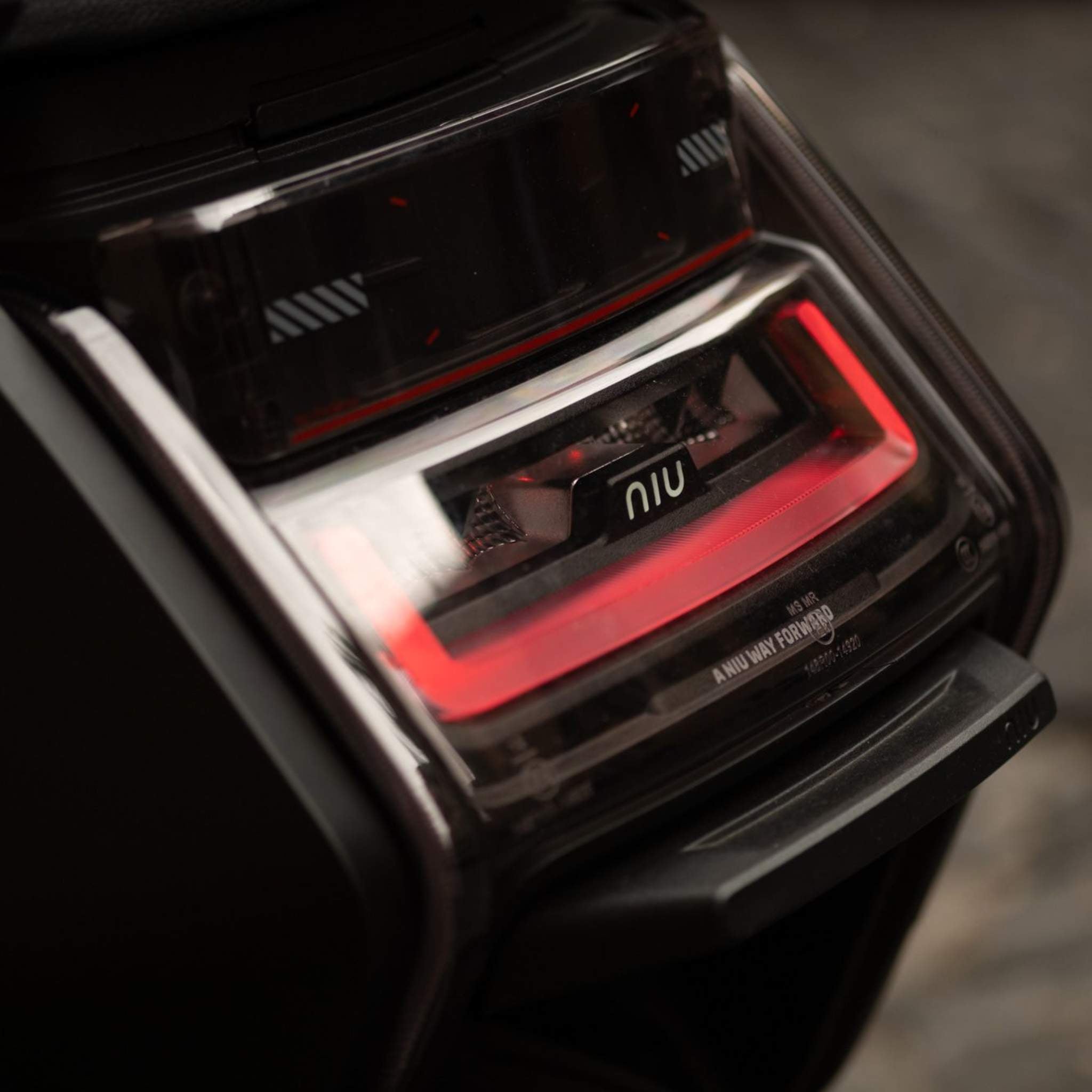 Nahaufnahme des Rücklichts des NIU Elektroroller E-Scooters Motorroller NQiX 500 mit auffälligen roten LEDs und dem NIU-Logo in der Mitte, in einem eleganten schwarzen und transparenten Gehäuse vor einem unscharfen Hintergrund.