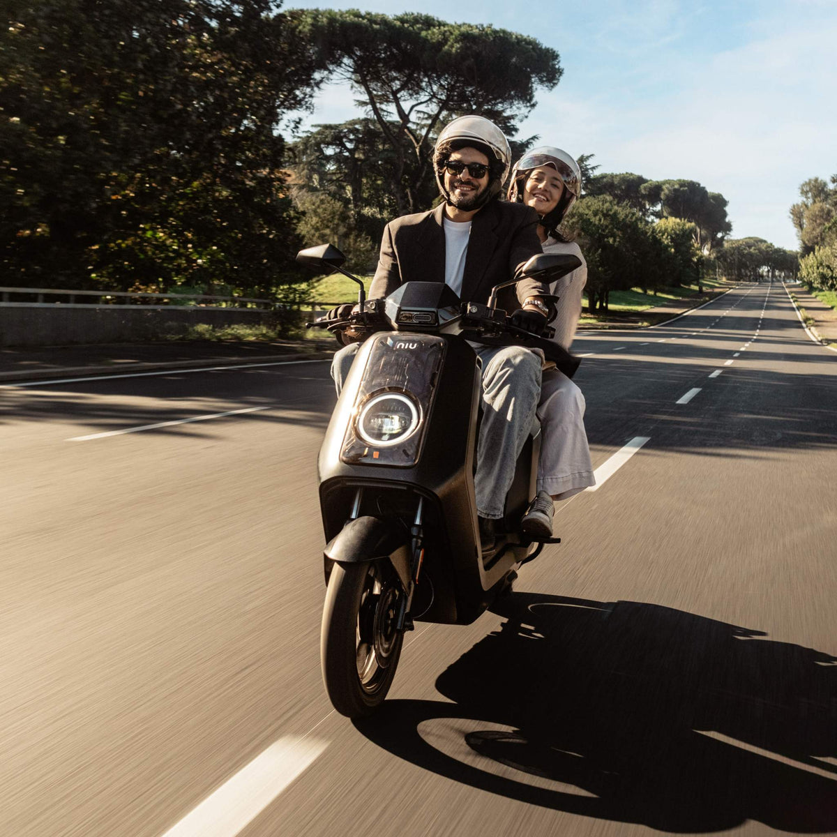 Zwei Personen mit Helmen fahren auf einem stylischen schwarzen NIU NQiX 500 Elektroroller (Modell 2025) mit einer Geschwindigkeit von bis zu 100 km/h eine sonnendurchflutete, von Bäumen gesäumte Straße entlang. Beide lächeln und genießen sichtlich ihre gemütliche gemeinsame Fahrt auf dem neuesten Elektroroller von NIU.