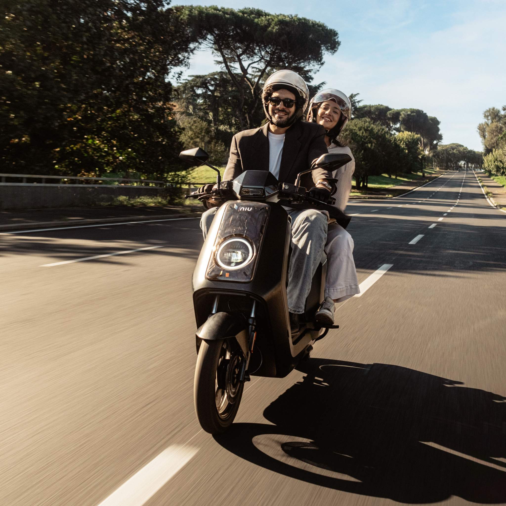 Zwei Personen mit Helmen fahren mit einem NIU Elektroroller Motorroller NQiX 500 (100 km/h) eine sonnige, von Bäumen gesäumte Straße hinunter. Beide lächeln und genießen eine sorglose Fahrt auf einer leeren Straße unter strahlend blauem Himmel.