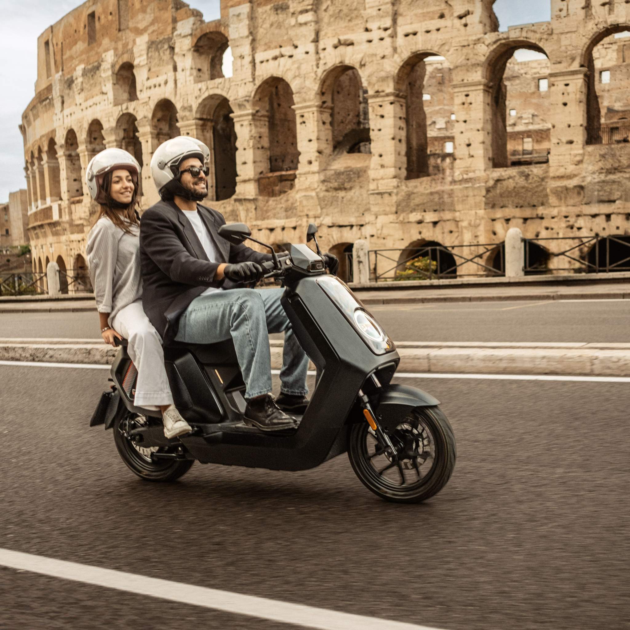 Zwei Personen mit Helmen fahren an einem sonnigen Tag mit einem stylischen Elektroroller NIU NQiX 500 (100 km/h) am Kolosseum in Rom vorbei - der Fahrer in schwarzer Jacke und Jeans, der lächelnde Beifahrer in heller Kleidung.