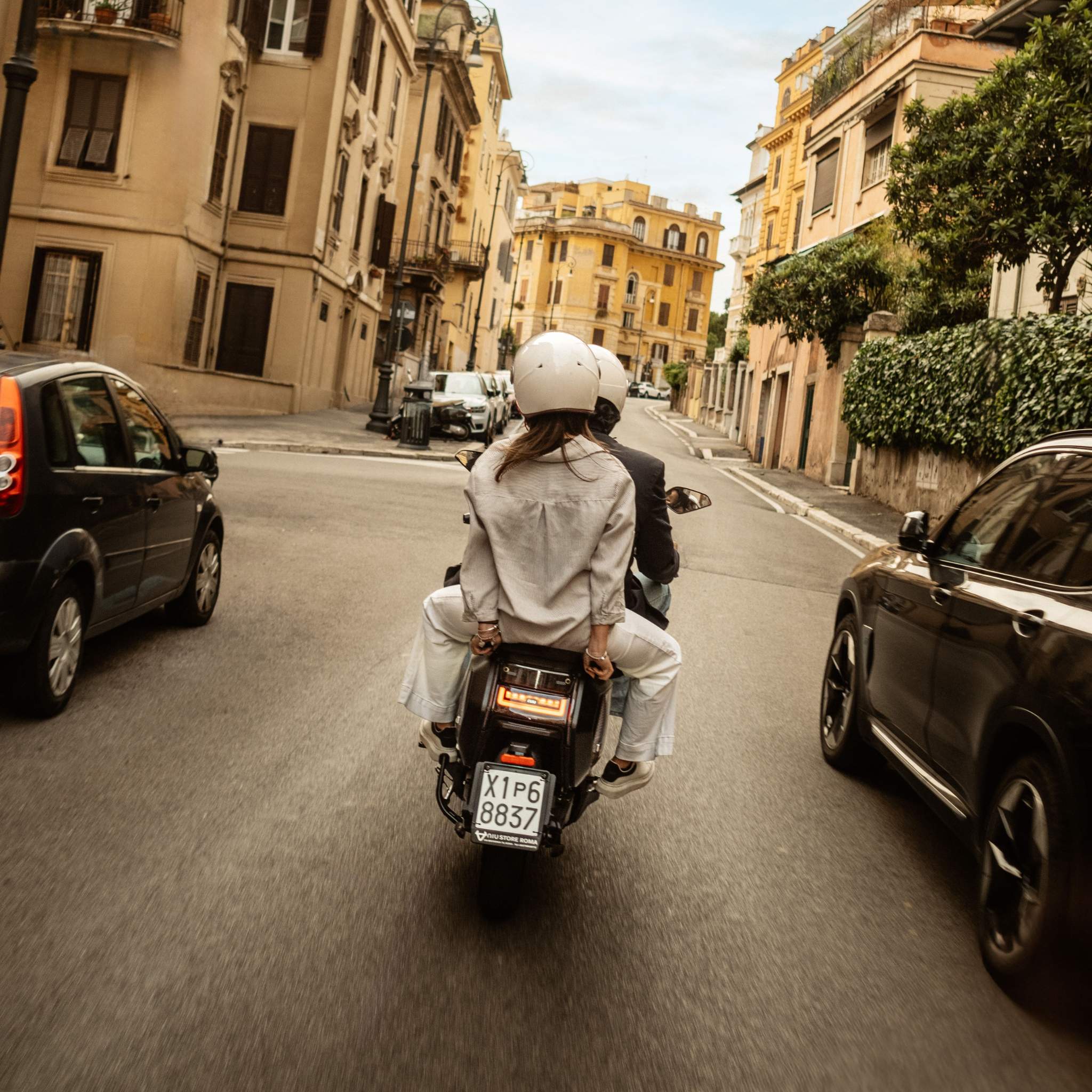 Zwei Personen mit Helmen fahren mit einem Elektroroller NIU NQiX 500 (100 km/h) an einem klaren Tag durch eine von parkenden Autos und historischen Gebäuden gesäumte Stadtstraße, von hinten gesehen. Die Straße ist relativ leer.