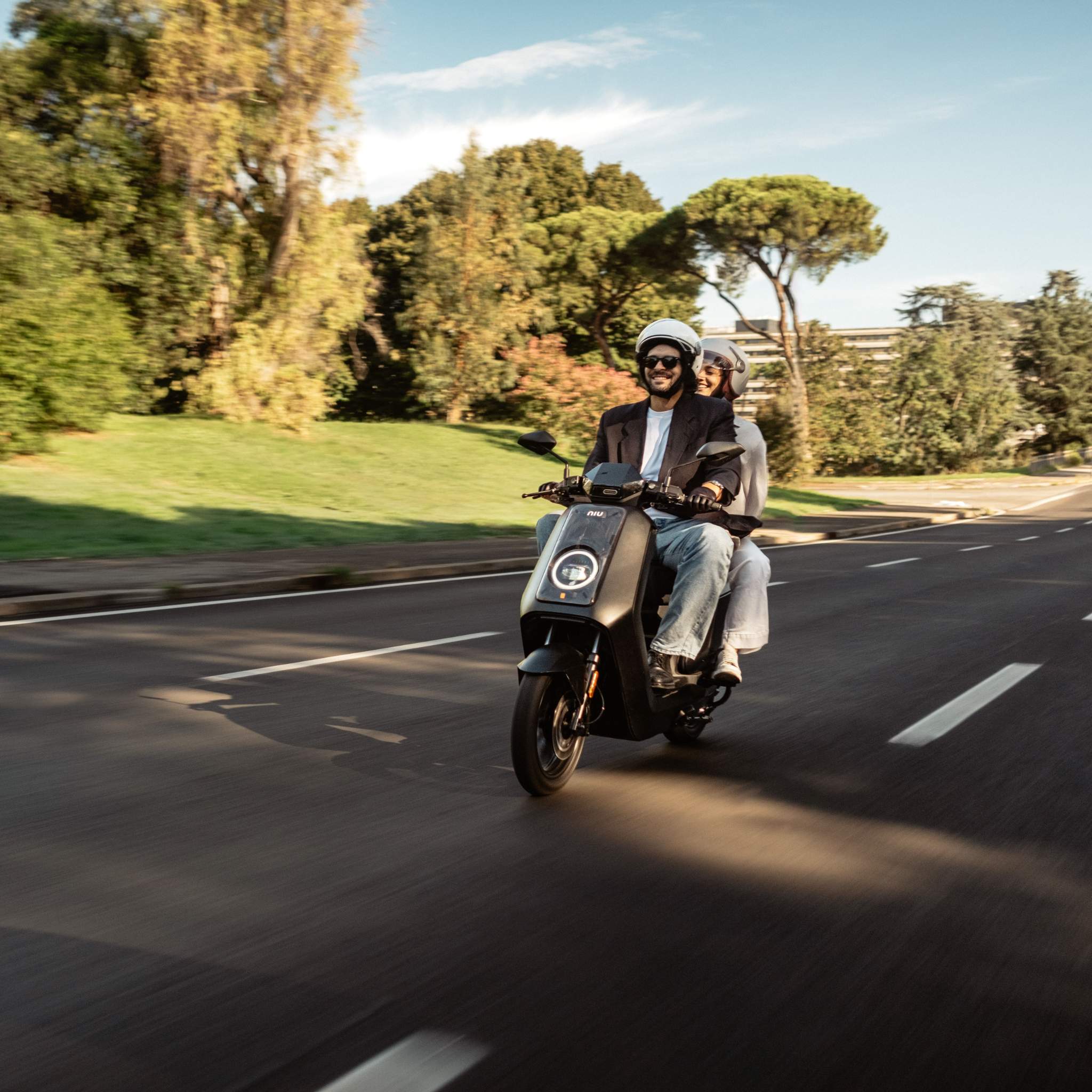 Zwei Personen mit Helmen fahren mit einem schwarzen NIU NQiX 500 Elektroroller Motorroller (100 km/h) auf einer breiten, leeren Straße, umgeben von Grünflächen und Bäumen, unter einem klaren Himmel.