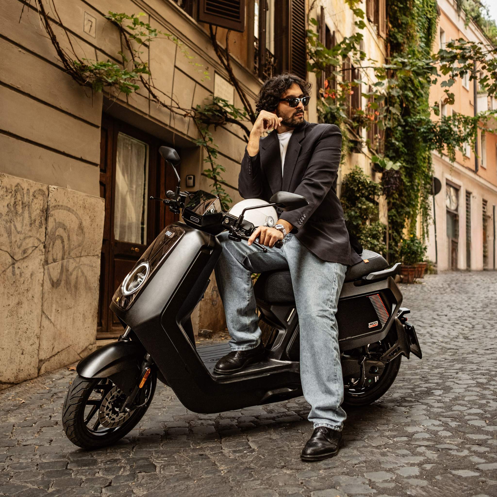 Ein Mann mit Sonnenbrille, dunklem Blazer und Jeans sitzt auf einem schwarzen NIU Elektroroller Motorroller NQiX 500 (100 km/h) auf einer Kopfsteinpflasterstraße mit alten, begrünten Gebäuden.