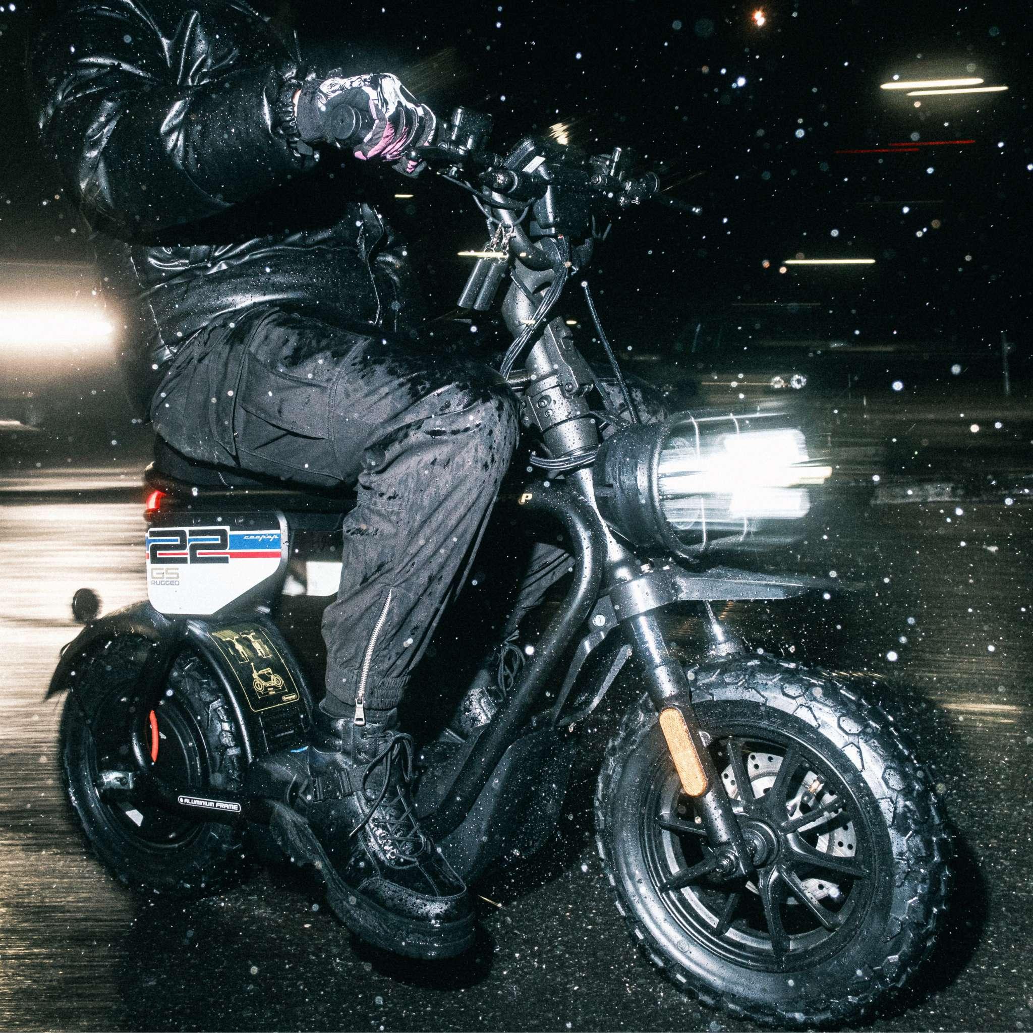 Eine Person in Schwarz fährt den Coopop E-Scooter E-Roller Rugged GS nachts im Regen, Wassertropfen sichtbar und verschwommene Lichter dahinter. Der Roller hat die Nummer 22 an der Seite und einen 1200-Watt-Nabenmotor.