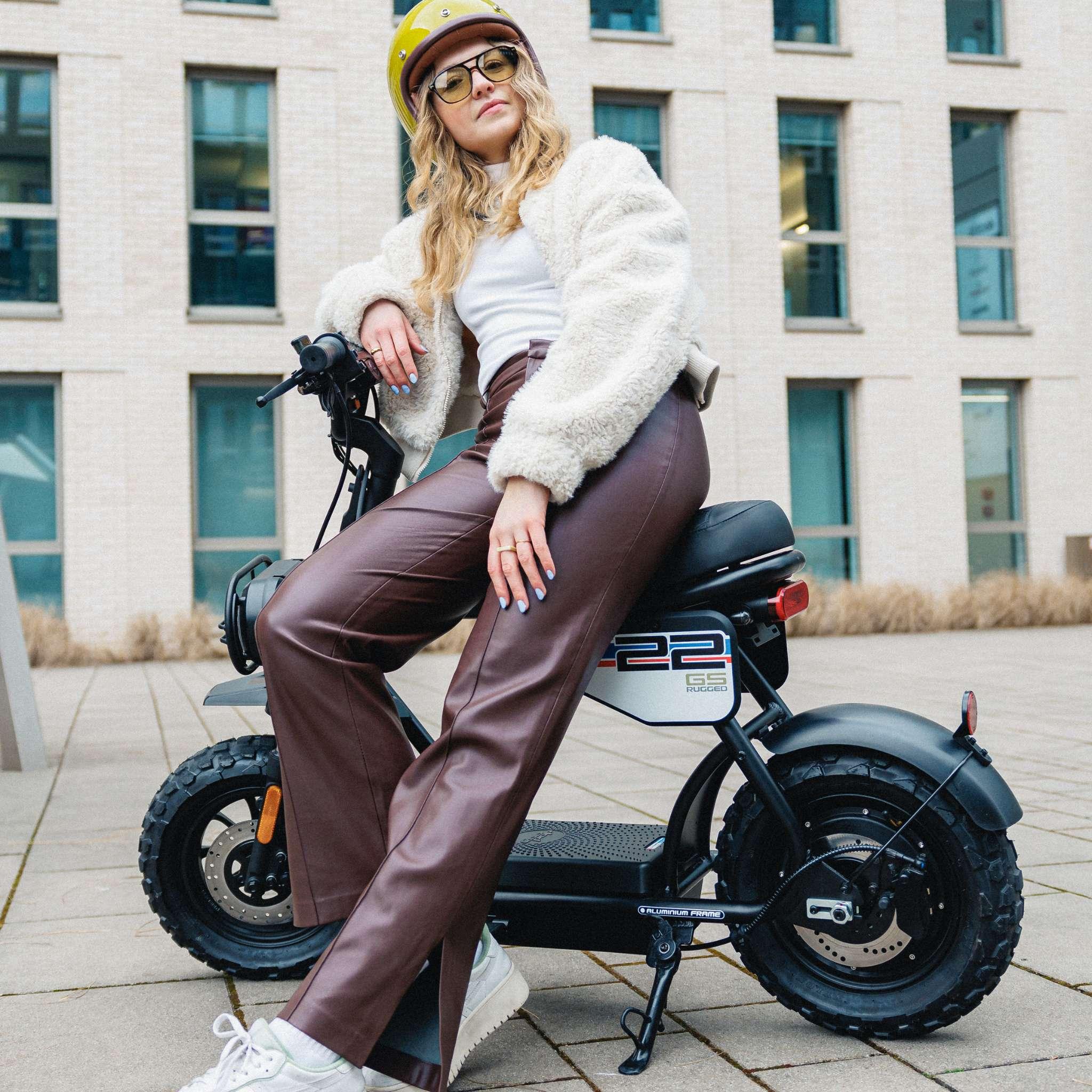 Eine Frau mit gelbem Helm, weißem Oberteil und brauner Lederhose sitzt selbstbewusst auf einem Coopop E-Scooter Rugged GS (45km/h & 25km/h) von Coopop, der mit einem 1200W Nabenmotor ausgestattet ist, und posiert im Freien vor einem modernen Gebäude mit großen Fenstern.