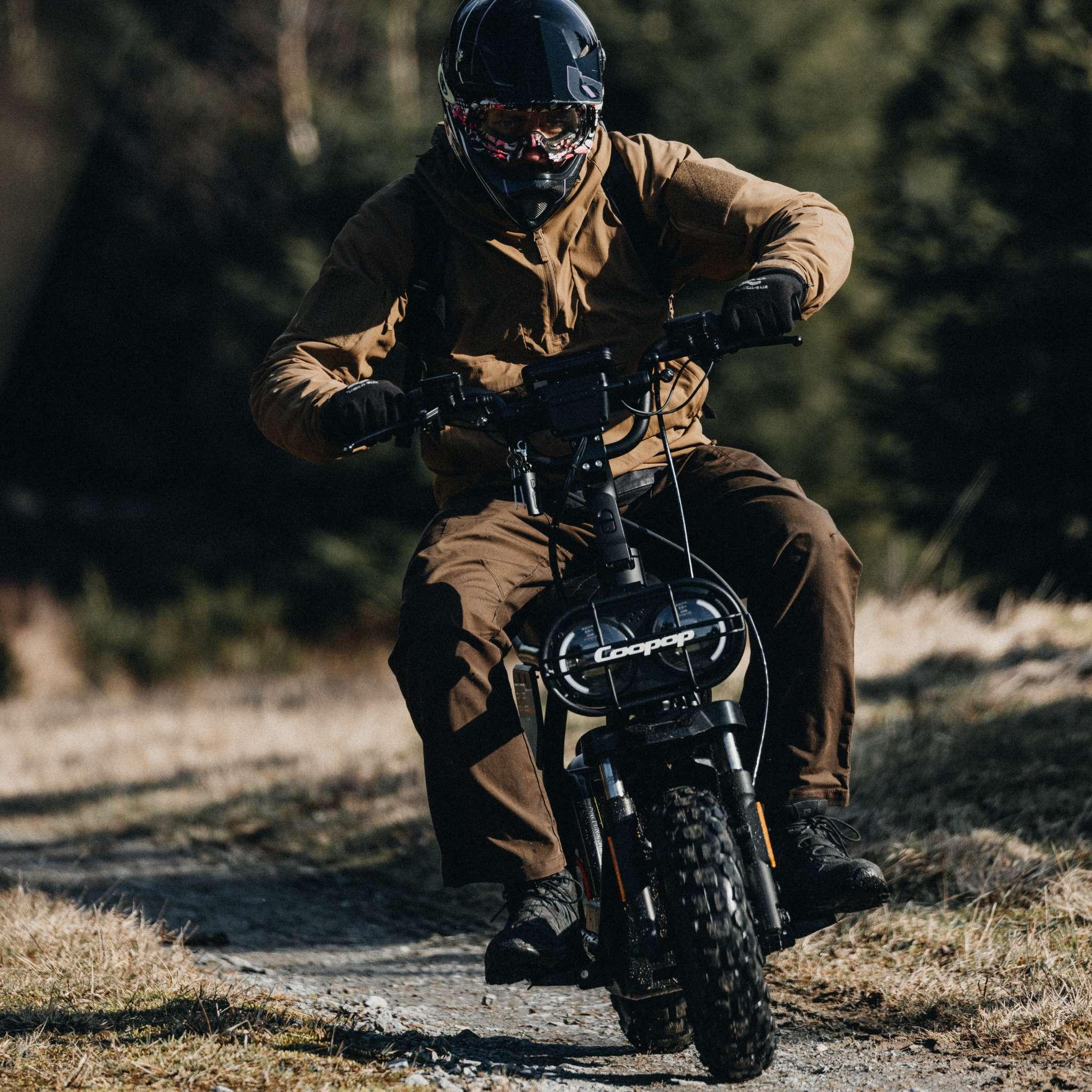Ein Fahrer in brauner Kleidung und mit Helm fährt auf dem Coopop E-Scooter E-Roller RUGGED GS (45km/h & 25 km/h) über einen schmalen Feldweg, während das Sonnenlicht durch die Bäume in einem grasbewachsenen Waldgebiet fällt.
