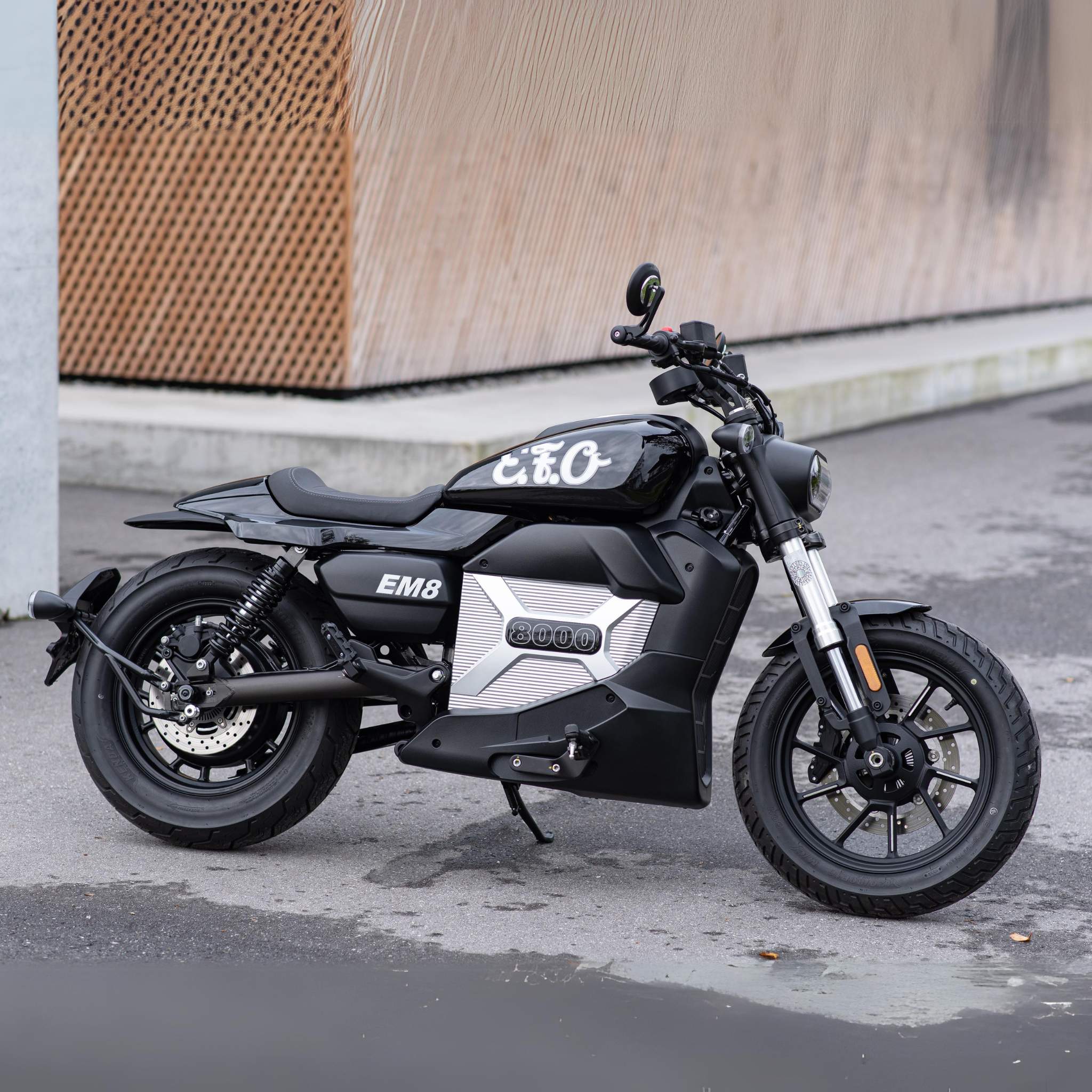 Ein schickes, schwarz-silbernes E.F.O. Elektro Motorrad EM8 120 km/h parkt vor einem modernen Gebäude mit braunen Paneelen und zeigt Innovation und Stil - und ist zudem förderfähig im Rahmen des Umweltprämie Aktionspreises!.