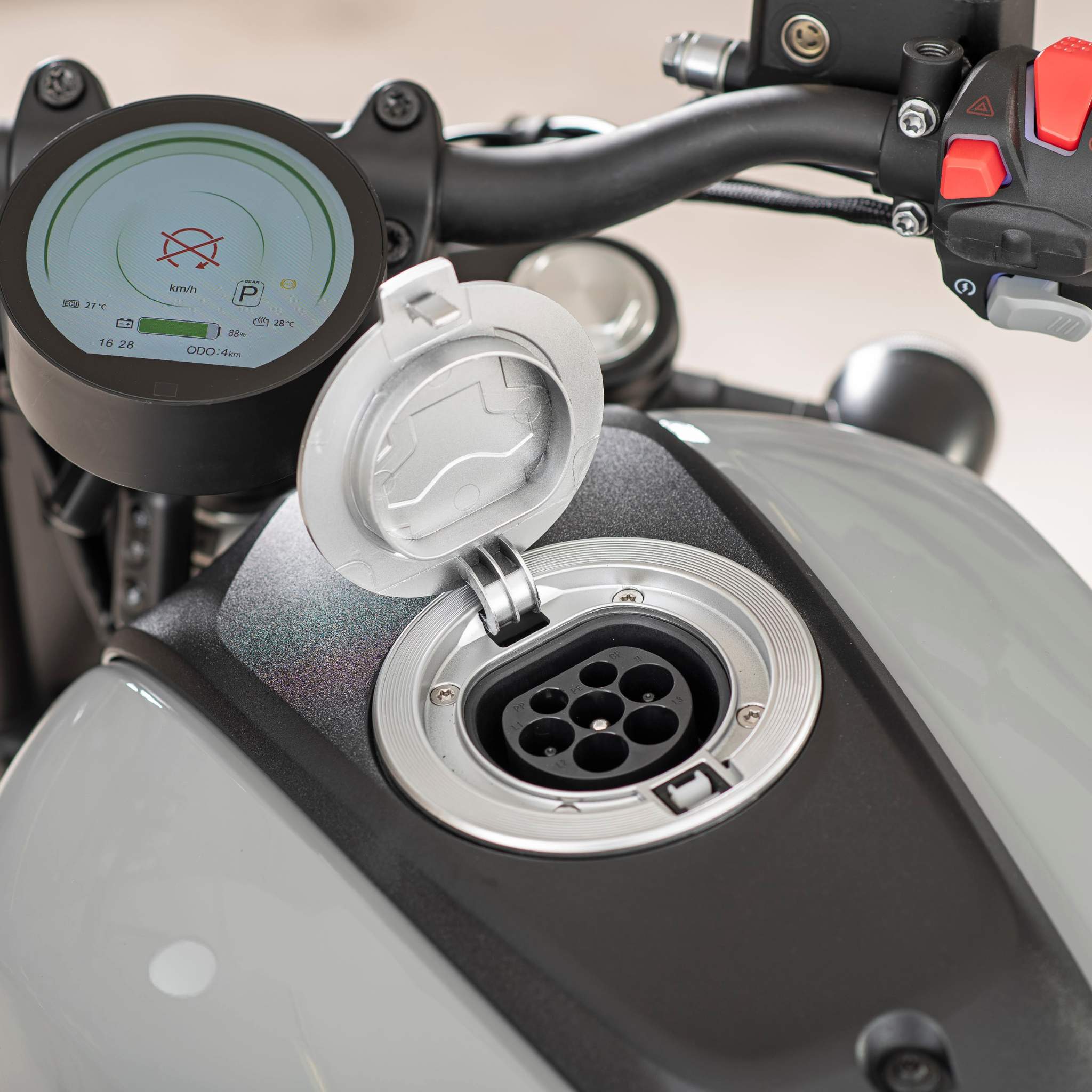 Nahaufnahme des Ladeanschlusses des Elektro Motorrad E.F.O EM8 120 km/h am Tank, Abdeckung geöffnet. Der Lenker und das digitale Display im Hintergrund zeigen Geschwindigkeit und Batterieinformationen an. Perfekt für Fahrer, die eine Umweltprämie suchen!.