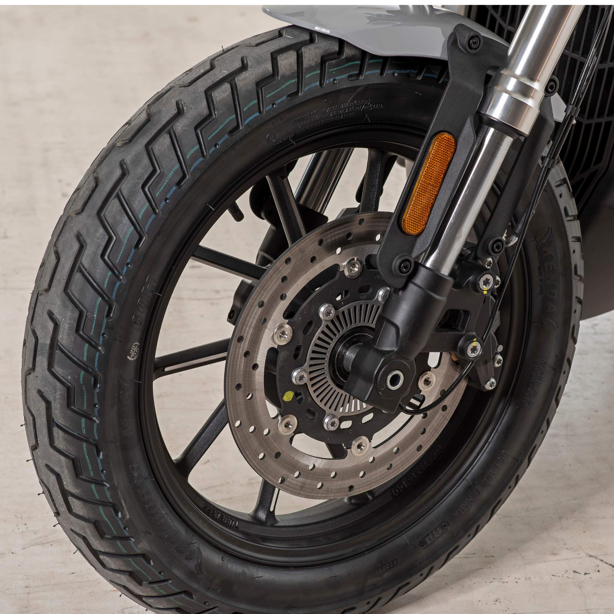 Nahaufnahme des Vorderrads eines E.F.O. Elektro Motorrad E.F.O EM8 120 km/h mit schwarzen Reifen, Scheibenbremse und Federgabel mit orangefarbenem Reflektor, geparkt auf hellem Beton. Förderfähig im Rahmen der Umweltprämie.