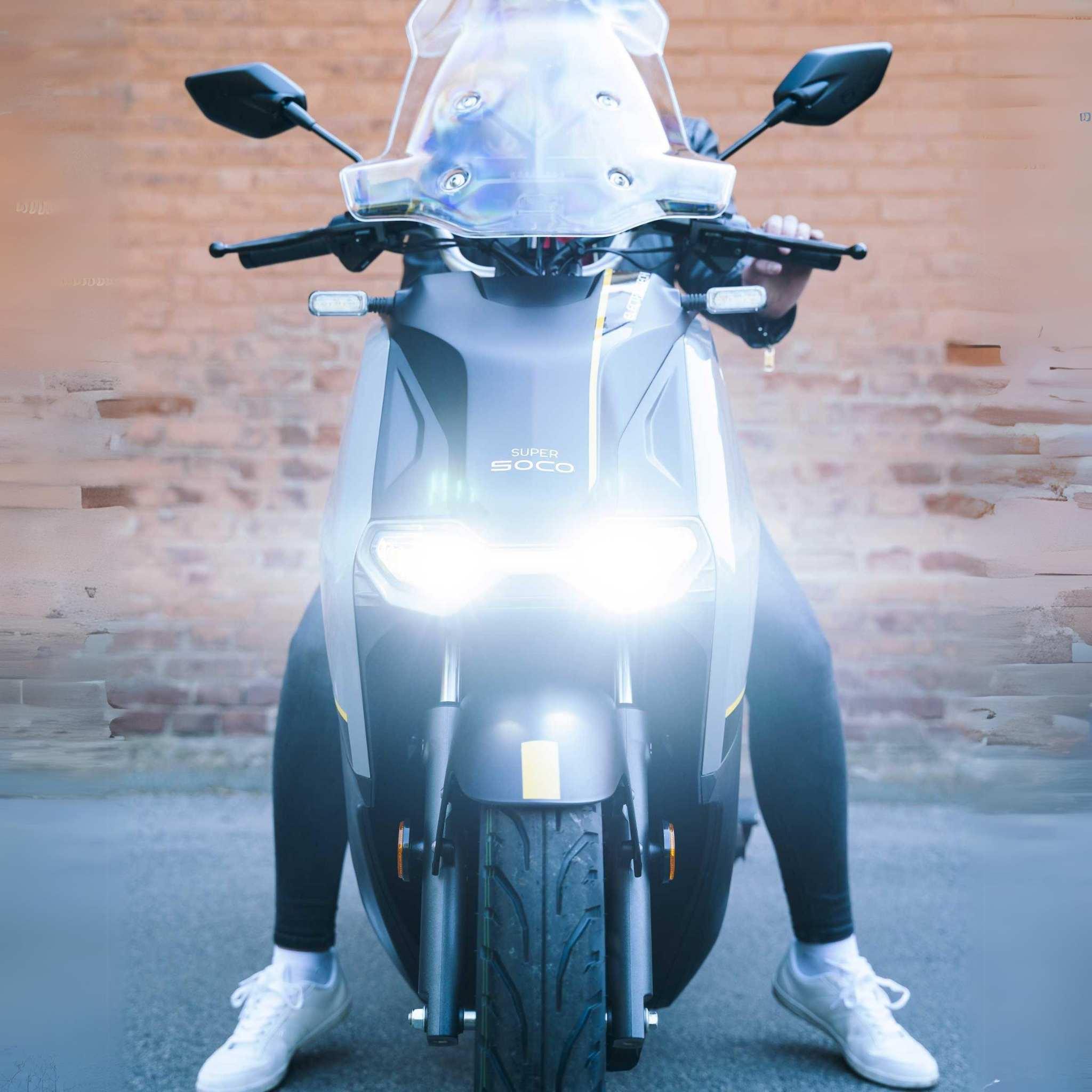 Ein Fahrer mit weißen Schuhen und dunkler Hose sitzt vorne auf einem Super Soco CPX Pro 105 km/h Elektroroller von Super Soco, die Scheinwerfer leuchten hell gegen eine unscharfe Backsteinwand. Der Roller erreicht eine Geschwindigkeit von bis zu 105 km/h.