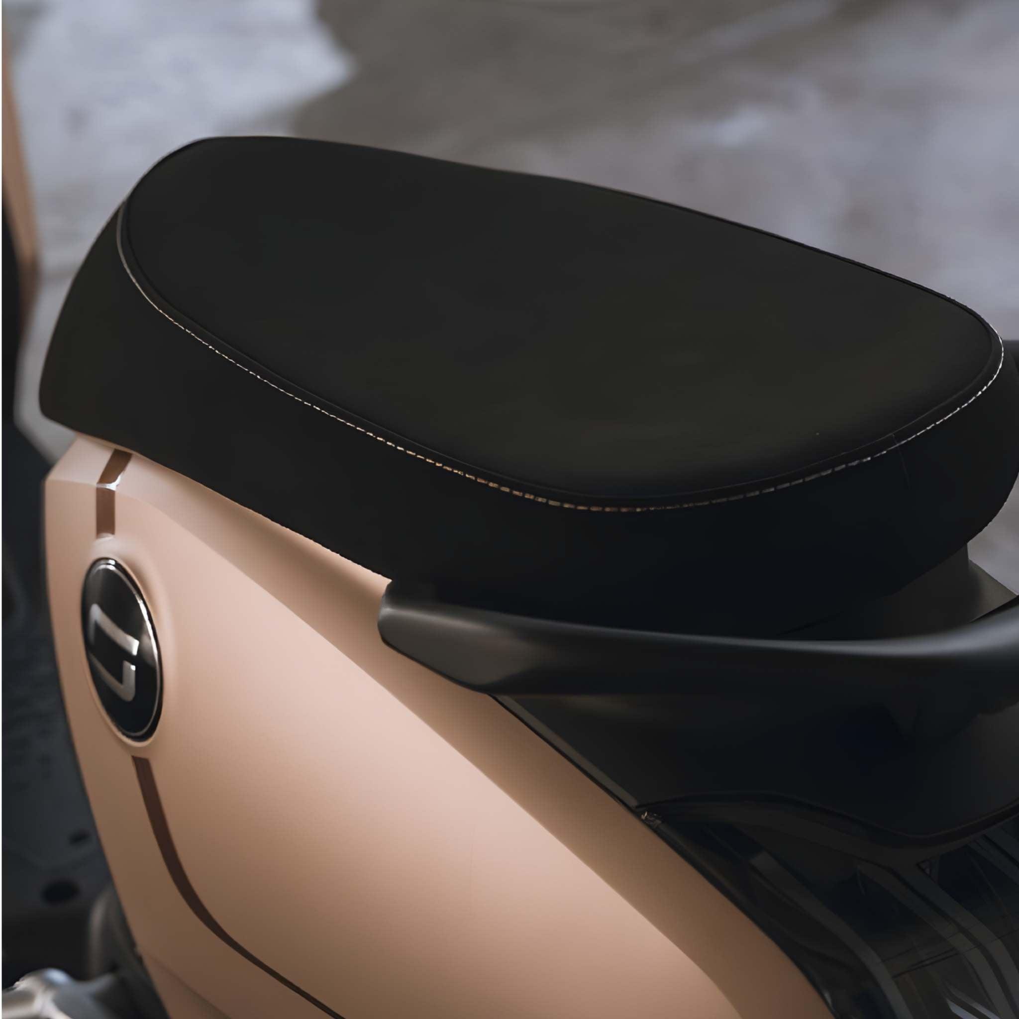 Nahaufnahme des schwarzen, gepolsterten Sitzes des Super Soco Elektrorollers CUx Pro mit beiger Karosserie und rundem Emblem. Der unscharfe Hintergrund unterstreicht das klare, moderne Design - ideal für umweltfreundliche Mobilität.