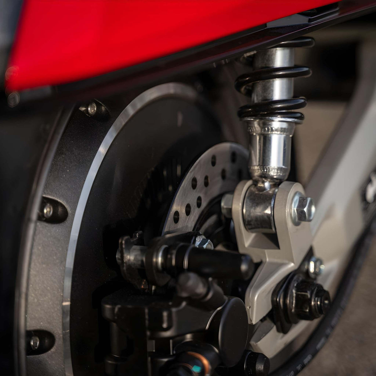 Nahaufnahme der Hinterradaufhängung des Super Soco E-Roller CUx Pro mit einem gewickelten Stoßdämpfer, einer Bremsscheibe und einer Schwinge unter einer roten Verkleidung, die die Technik hinter der umweltfreundlichen Mobilität hervorhebt.