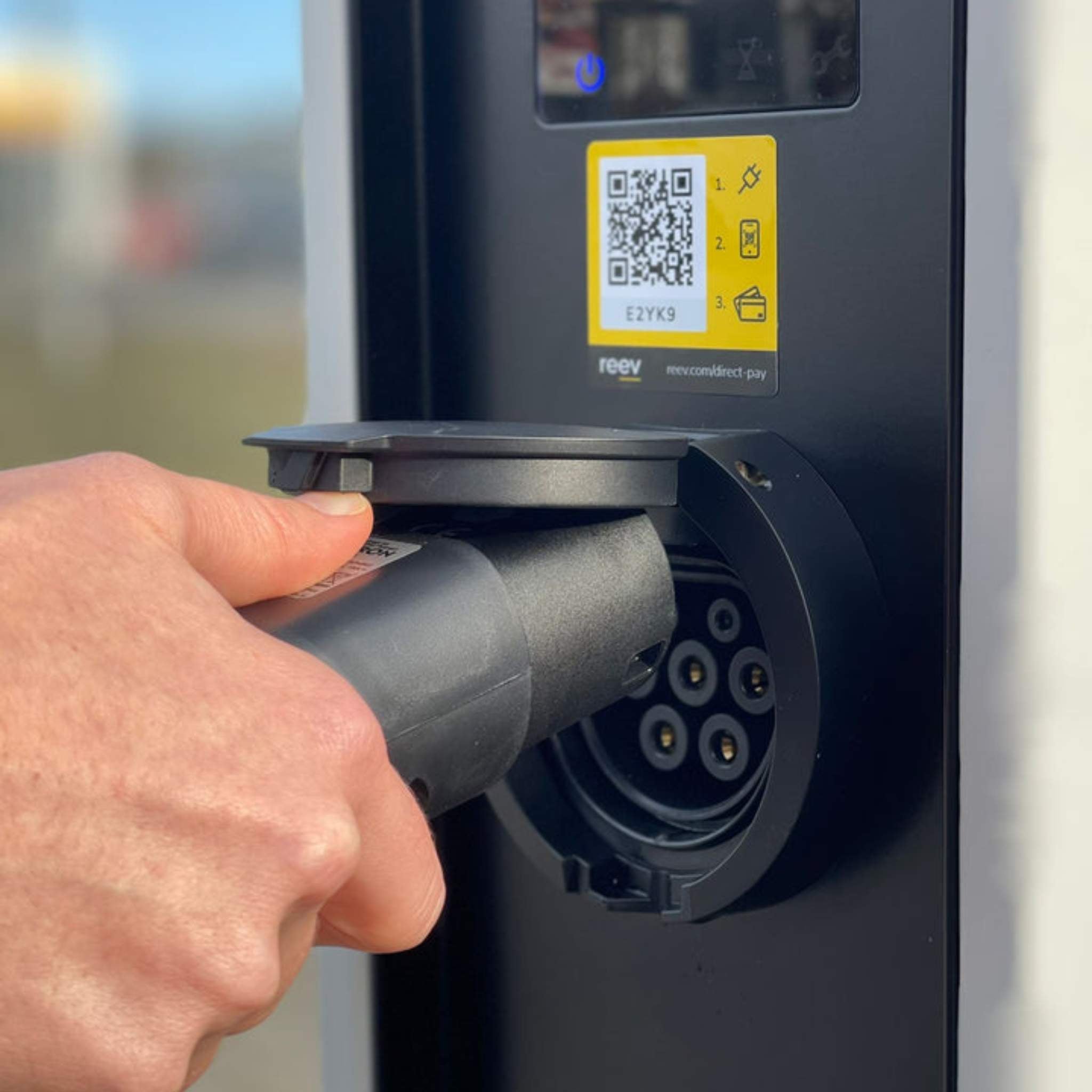 Eine Hand steckt ein Ladekabel mit dem TYP2 Adapter für Schuko Stecker von VMOTO in eine Ladesäule, um ein Elektro Motorrad zu laden. Ein QR-Code, eine Bezahlanleitung und der Adapter sind auf dem Gerät sichtbar.