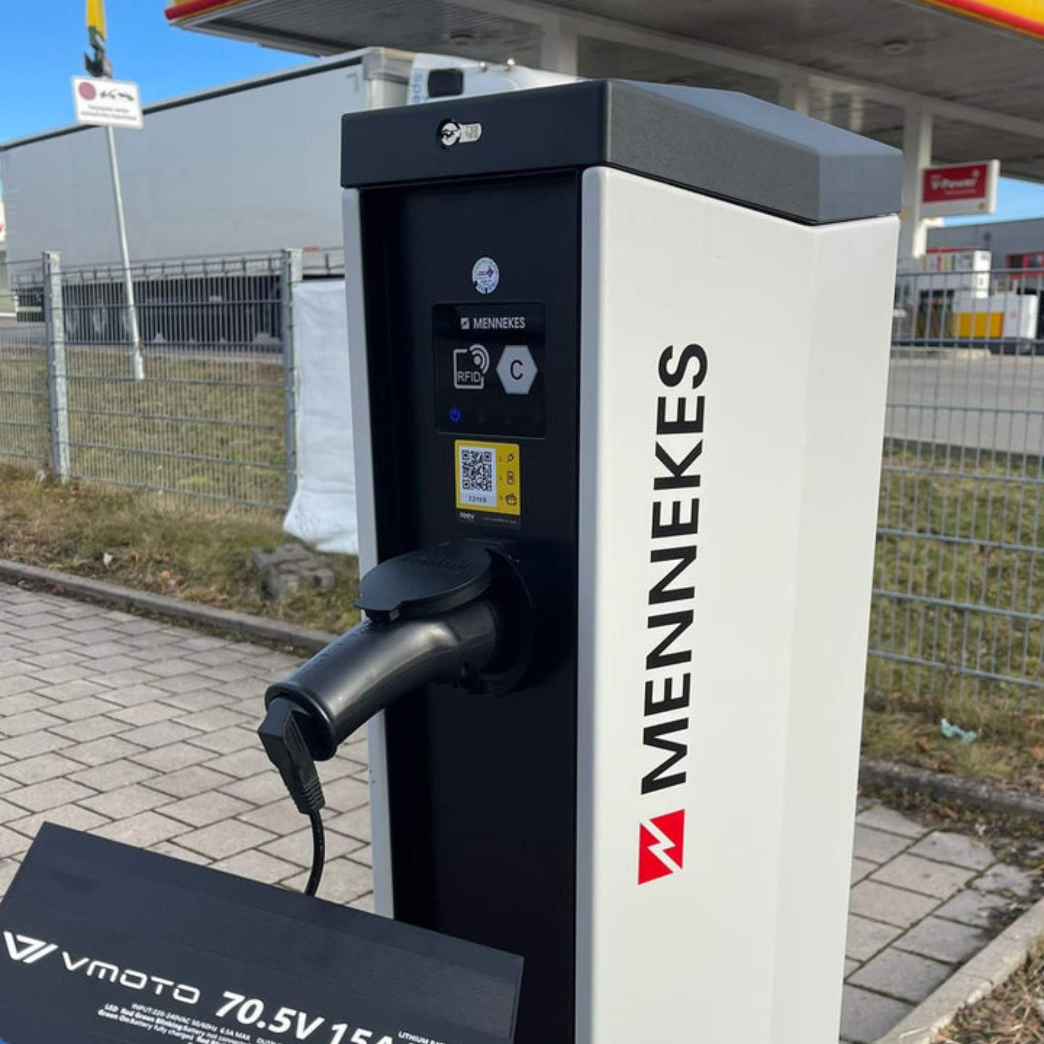 Ein VMOTO TYP2 Adapter für Schuko Stecker - die zuverlässige Ladelösung für dein Elektro Motorrad, E-Chopper, E-Roller & mehr - liegt an einer Ladesäule auf Pflaster nahe einer Tankstelle. Im Hintergrund sind Gras und ein Zaun zu sehen.