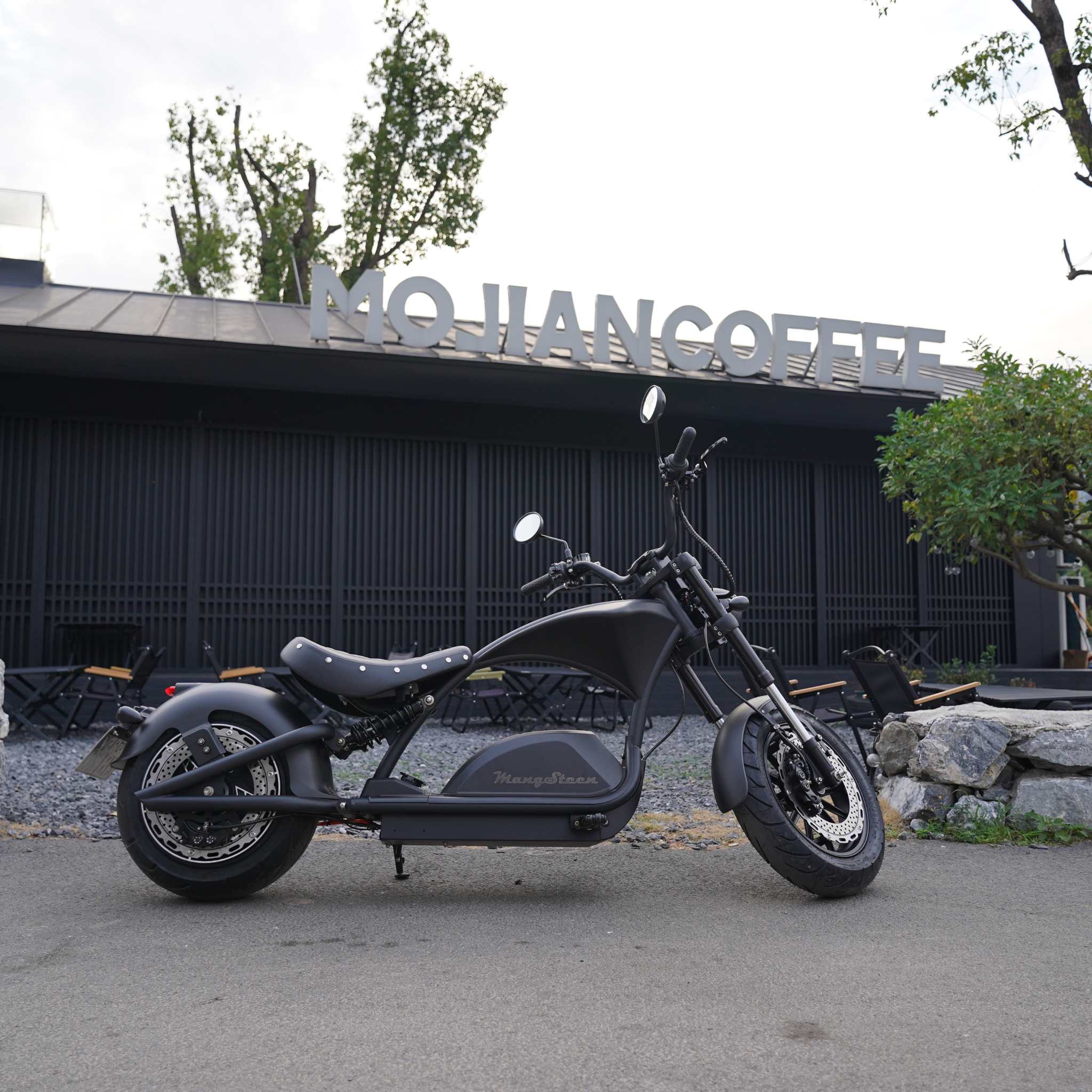Ein Mangosteen E-Chopper M1PS (85 km/h, 40 Ah Samsung Lithium-Akku, 4000 Watt) steht vor einem modernen Café mit MO JIAN COFFEE-Schild, umgeben von Bäumen, leeren Stühlen und einem Steingarten - ein Beispiel für urbanen Elektromotorrad-Stil.