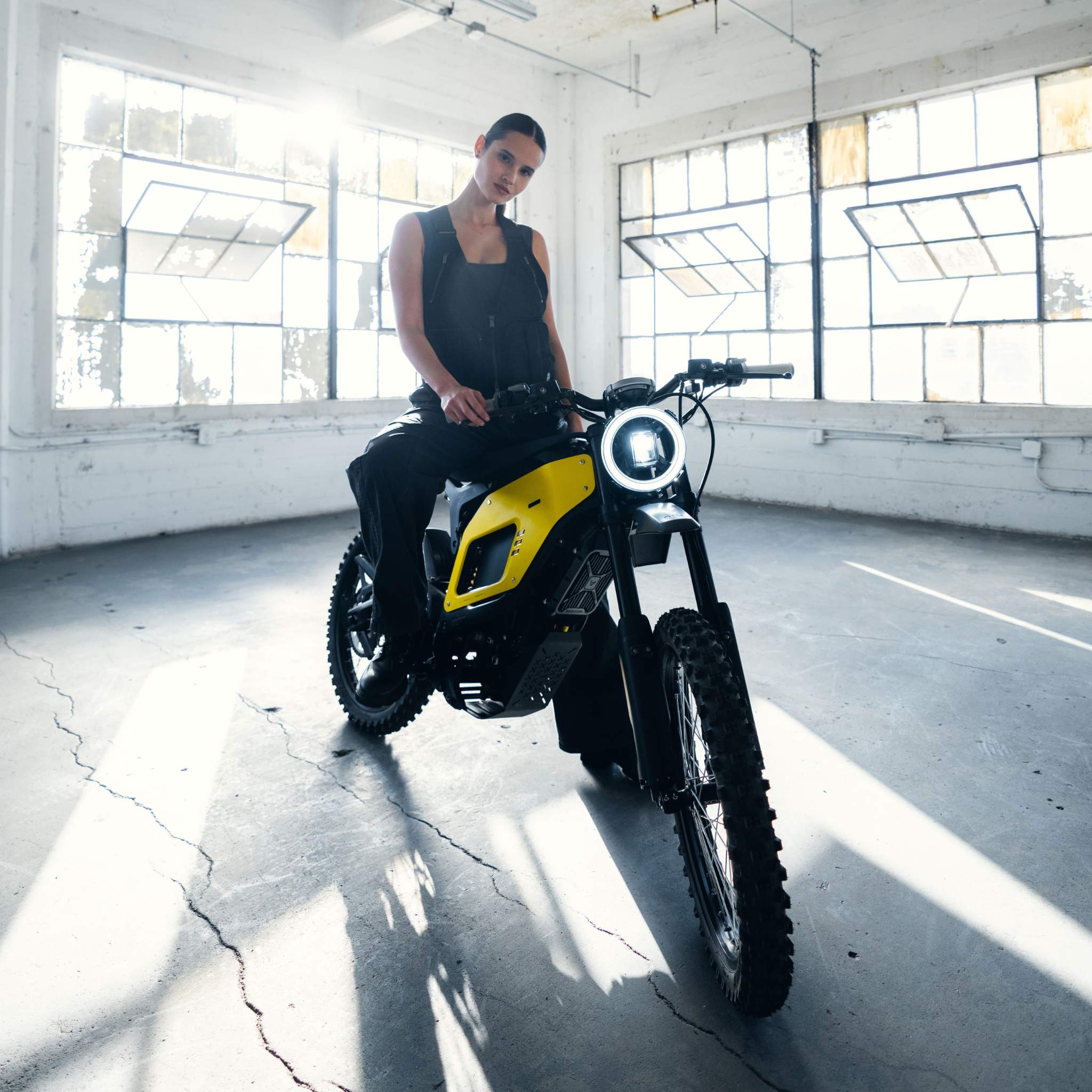 Eine dunkel gekleidete Person sitzt selbstbewusst auf einem NIU XQi3 Enduro Dirtbike Cross Motorrad (45 km/h) von NIU in einer sonnenbeschienenen Industriehalle mit großen Fenstern und Betonboden.