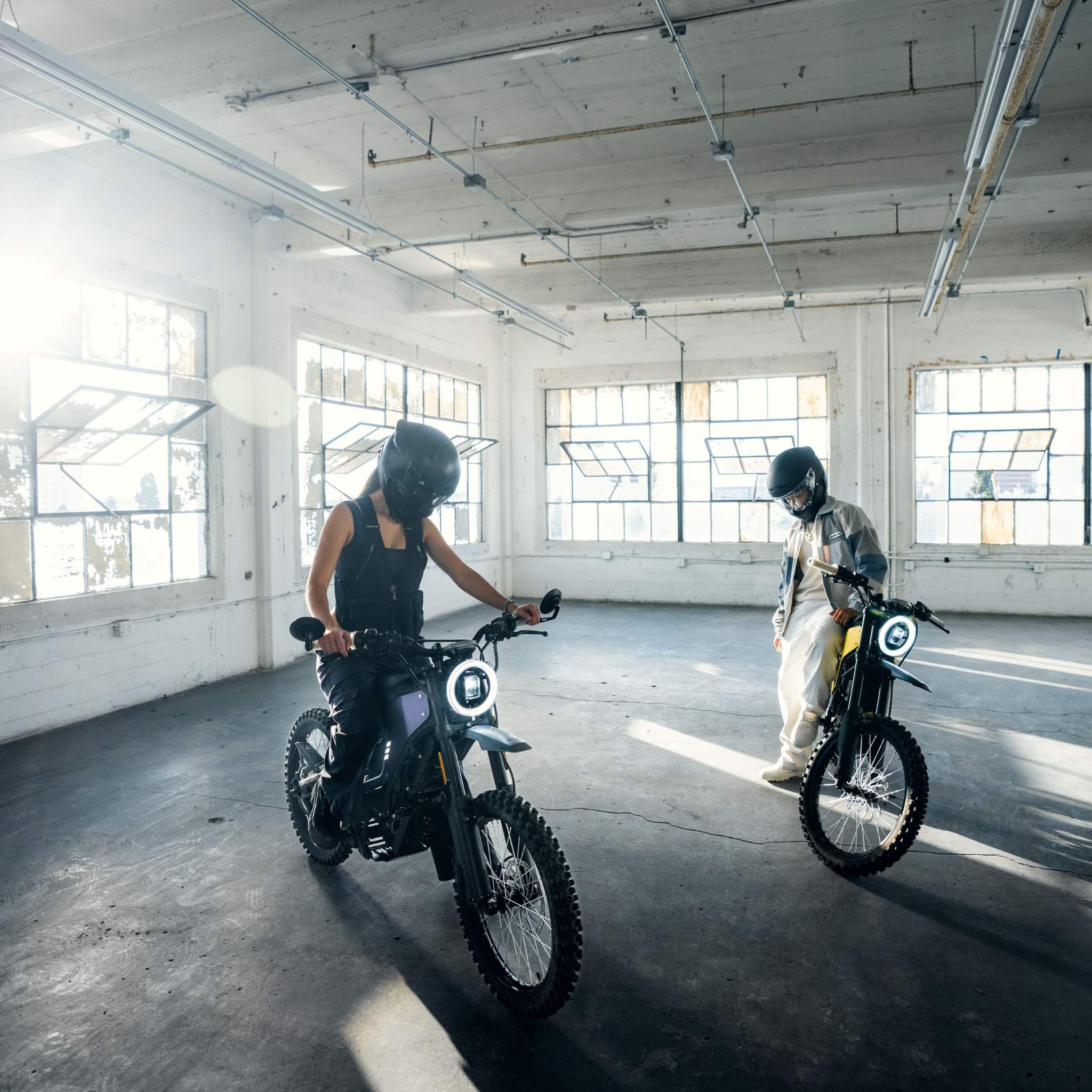Zwei Personen mit Helmen stehen in einem sonnendurchfluteten Industrieraum mit großen Fenstern und bereiten sich darauf vor, ihr NIU XQi3 Enduro Dirtbike Cross Motorrad 45 km/h von NIU zu fahren oder zu überprüfen.
