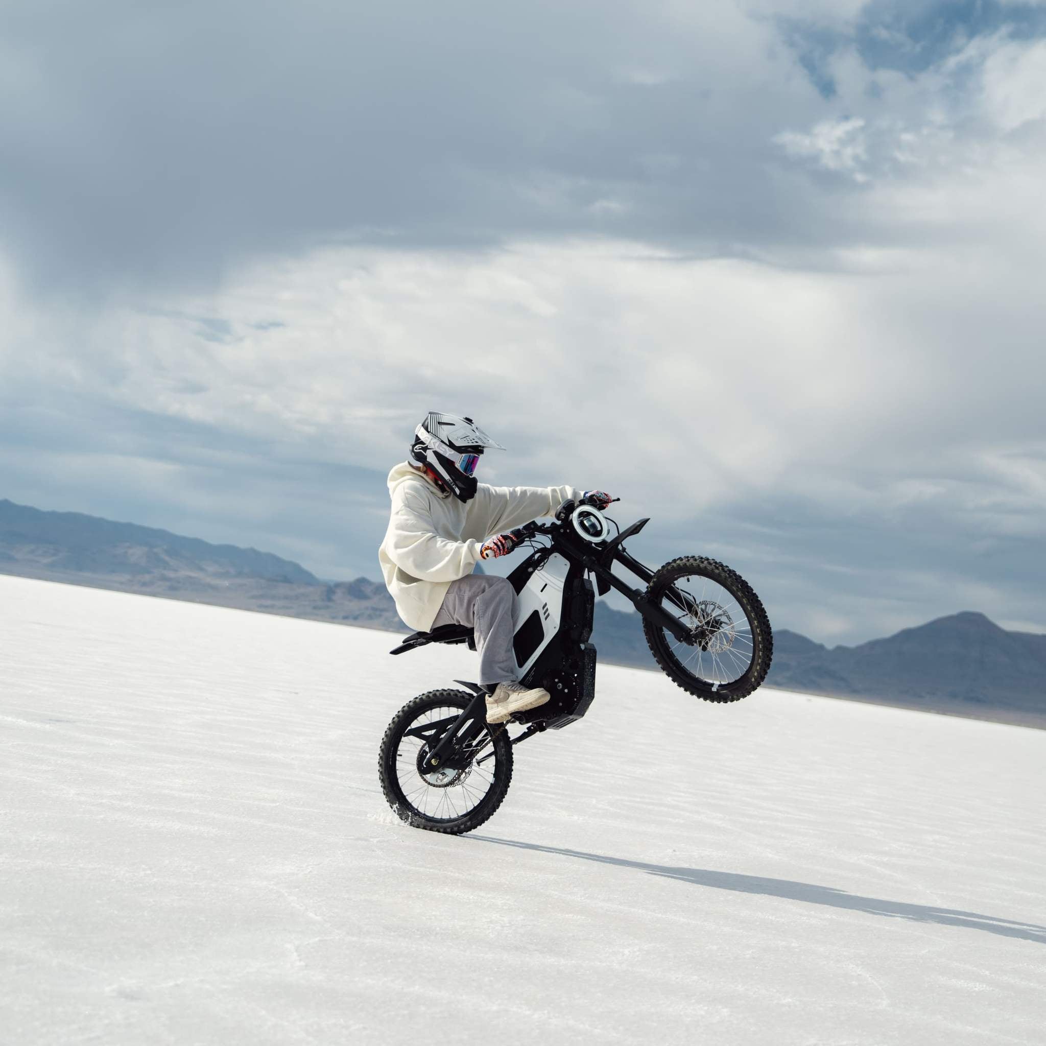 Ein Motorradfahrer mit Helm und weißem Kapuzenpulli macht einen Wheelie auf einem NIU XQi3 Enduro Dirtbike Cross Motorrad mit 45 km/h und rast durch die weiße Salzwüste mit Bergen und Wolken im Hintergrund.