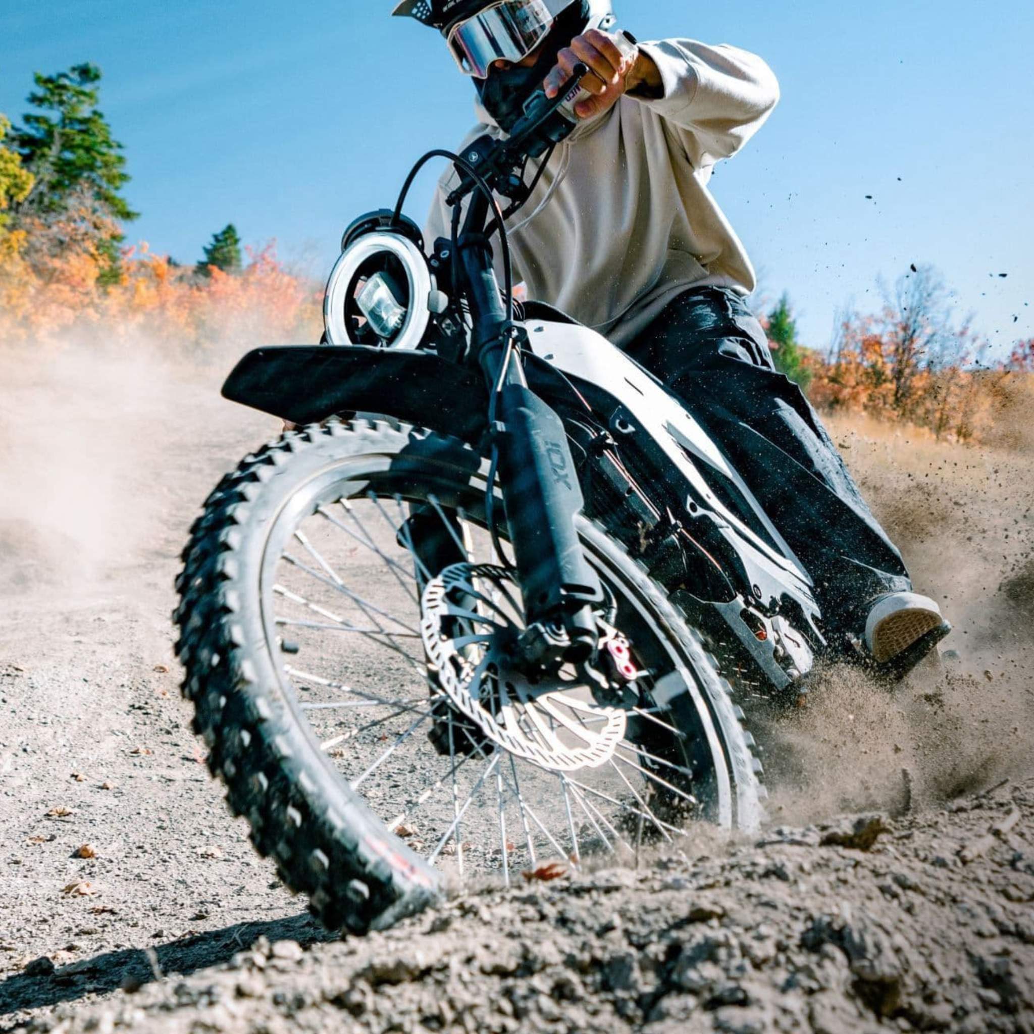Ein Fahrer mit Helm fährt auf einem NIU XQi3 Enduro Dirtbike Cross Motorrad mit 45 km/h um eine Kurve und wirbelt an einem sonnigen Tag mit Herbstbäumen im Hintergrund Staub auf.