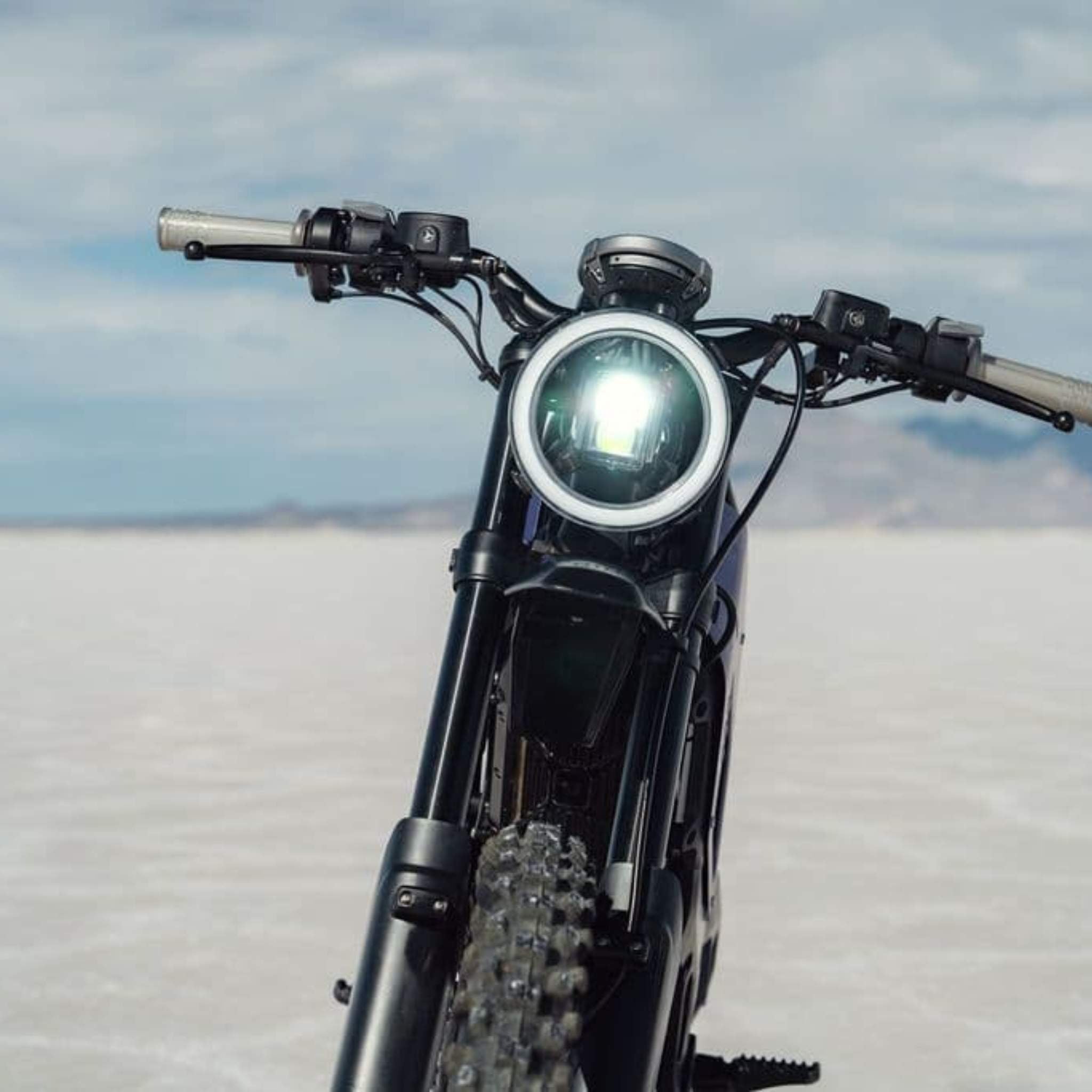Frontansicht des NIU XQi3 Enduro Dirtbike Cross Motorrad 45 km/h von NIU mit einem hellen, runden Scheinwerfer, dargestellt im Freien auf einer flachen, weißen Fläche unter einem bewölkten Himmel.