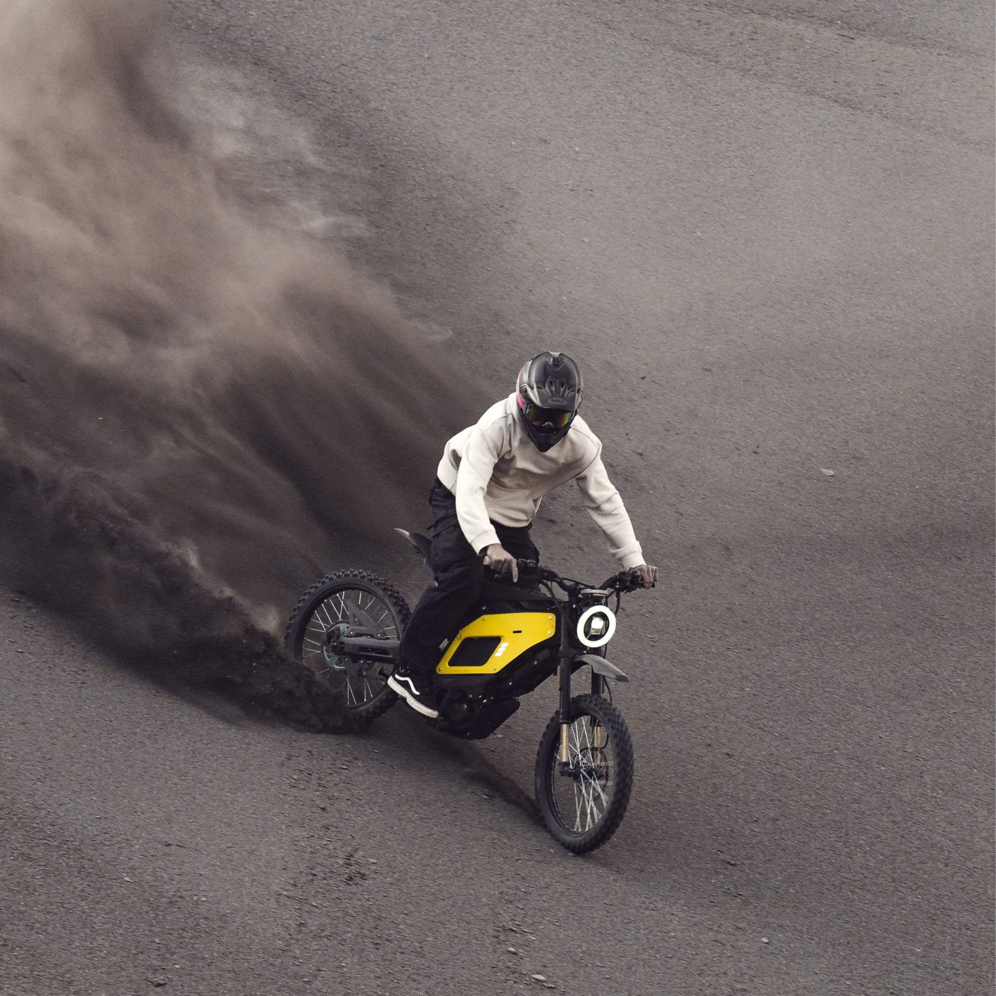 Eine Person mit Helm und leichter Jacke fährt auf einem gelben NIU XQi3 Enduro Dirtbike Cross Motorrad 45 km/h by NIU einen steilen, sandigen Abhang hinunter und wirbelt dabei eine große Spur dunklen Staubs auf.